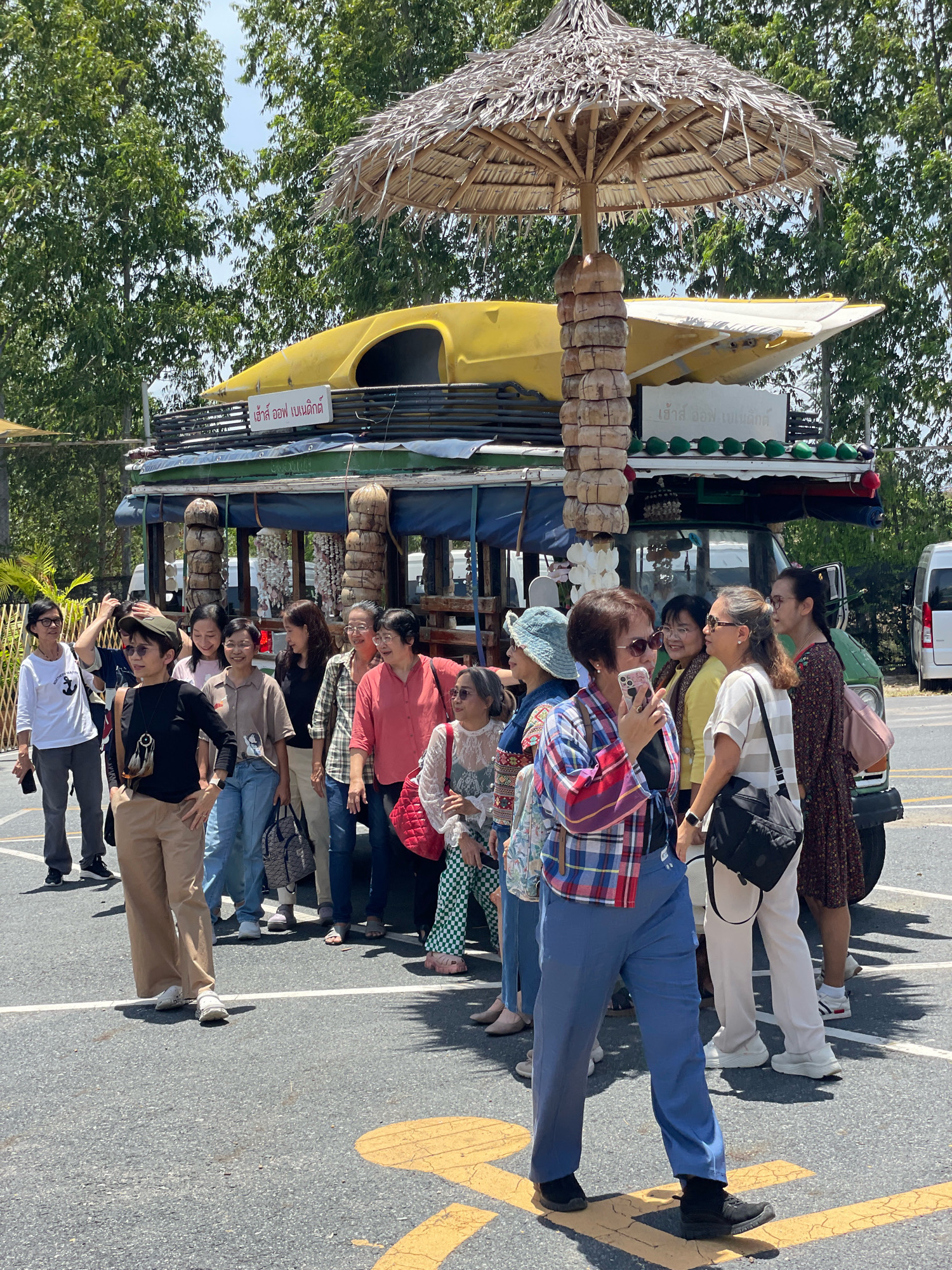 In Pattaya City, Thailand, a group of women poses for a photograph in front of a yellow boat-like structure mounted on a vehicle. The vehicle appears to be a modified truck, adorned with a thatched umbrella for shade and decorated with coconuts. The women are of various ages and are dressed in casual attire, some holding bags or taking pictures with their phones. The ground is asphalt with yellow markings, suggesting it might be a parking lot or a public area. In the background, lush green trees and a clear sky are visible, indicating a sunny day.