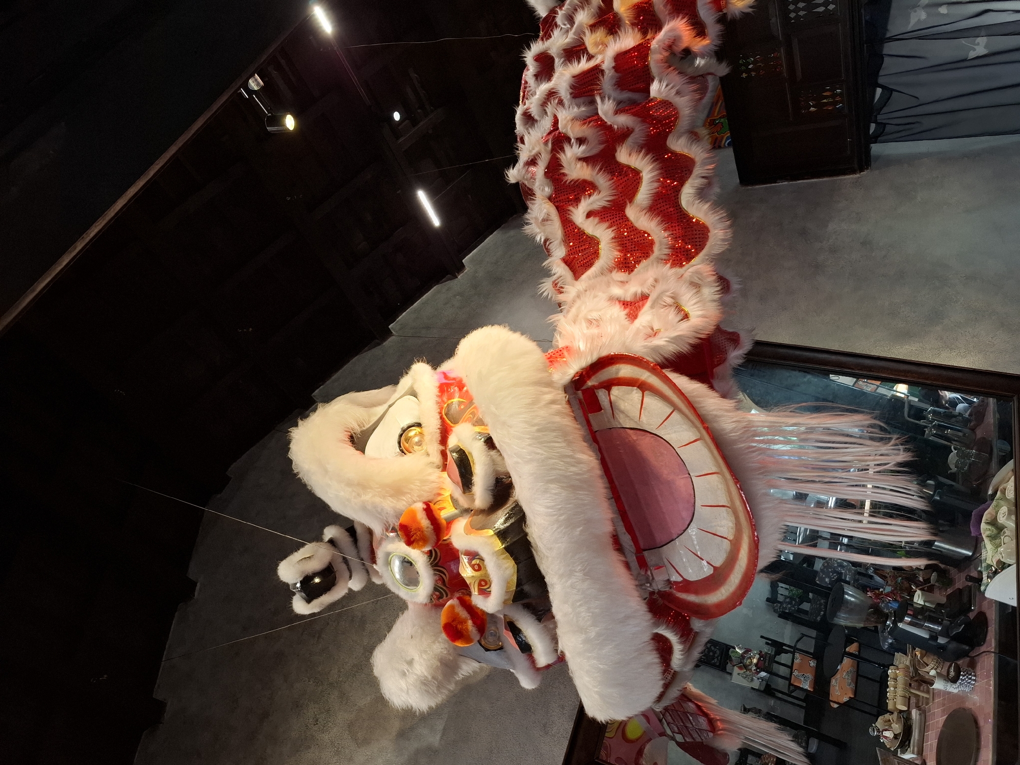 The image shows a detailed view of a vibrantly colored lion dance costume, likely displayed indoors. The lion's head, predominantly white and red with ornate golden accents and red pom-poms, hangs from the ceiling. Its body, a segmented structure covered in shimmering red sequins and edged with white faux fur, extends to the right. The backdrop includes a dark ceiling with industrial lighting fixtures, and a grey textured wall. Below, a reflection in a glass surface reveals a domestic or commercial setting with tables, chairs, and various objects, including a coffee machine and decorative items. The location is specified as Nha Trang Ward, Vietnam. There are no people clearly visible within the frame of the lion dance costume or its immediate surroundings.