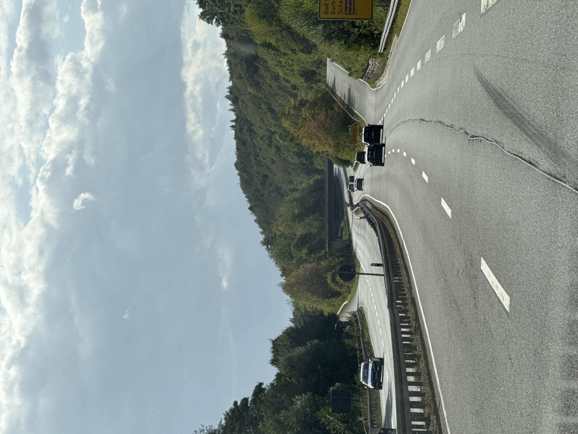 The image shows a highway in a rural, wooded area. The road curves through rolling hills covered in dense green trees. Several cars are visible on the highway, traveling in the same direction. The sky is overcast with scattered clouds. A yellow sign is visible on the right side of the road, displaying text that appears to be German. Based on the sign, the location is identified as Bad Lauterberg im Harz, Germany, with destinations including Bad Grund, Scharzfeld, and Bad Sachsa. The highway appears to be a multi-lane road with clear lane markings and guardrails. There is a noticeable crack running down the center of the road in the foreground.