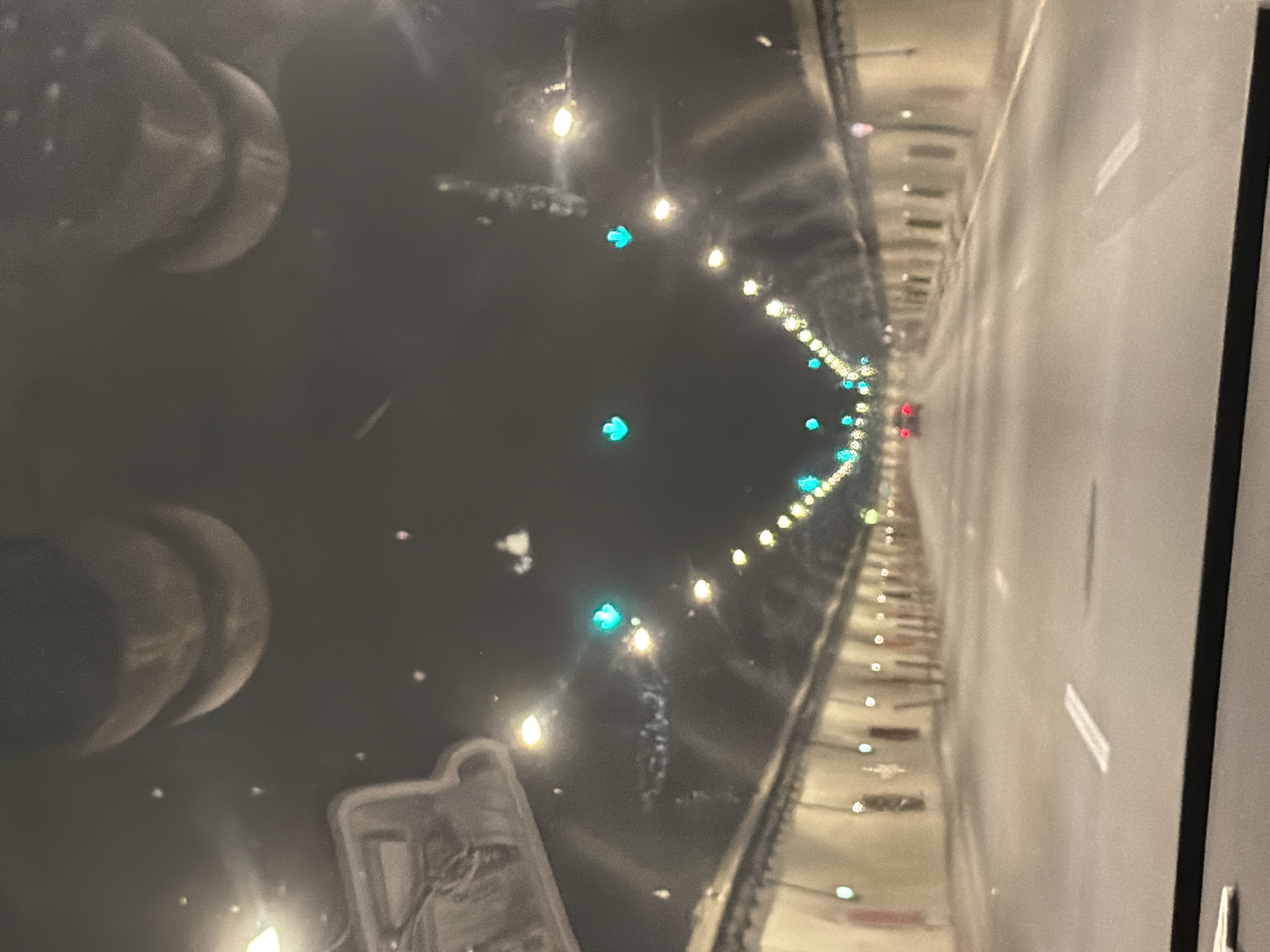 The image shows the interior of a tunnel, likely a highway tunnel, at night. The perspective is from inside a vehicle looking forward. The tunnel walls are lined with lights, some of which appear to be green directional lights, and others are white or yellow. There are also some red markings on the walls, possibly indicating emergency exits or equipment. The road ahead is paved and marked with white lines. Two cars are visible in the distance, their brake lights illuminated. The ceiling of the tunnel is dark, with some lights and structural elements visible. The overall impression is one of a functional, modern tunnel.
There is no discernible visual information in the image that would associate it with the city of Gorno Kamartsi, Bulgaria.