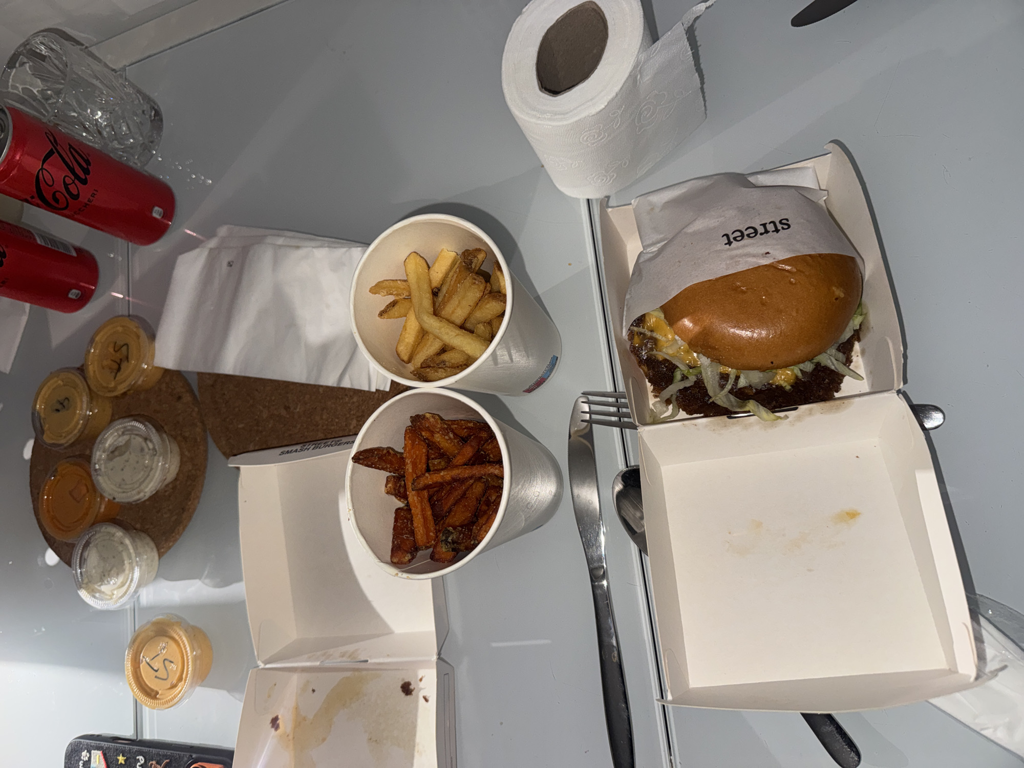 The image shows a meal on a gray surface. The meal includes a burger in a white cardboard box with the word "street" printed on the wrapper, two white paper cups containing french fries and sweet potato fries, an assortment of dipping sauces on a round cork coaster, and two cans of Coca-Cola. Also present is a roll of toilet paper and a set of silverware consisting of a fork and a spoon. There is no indication of the location or any people in the image.