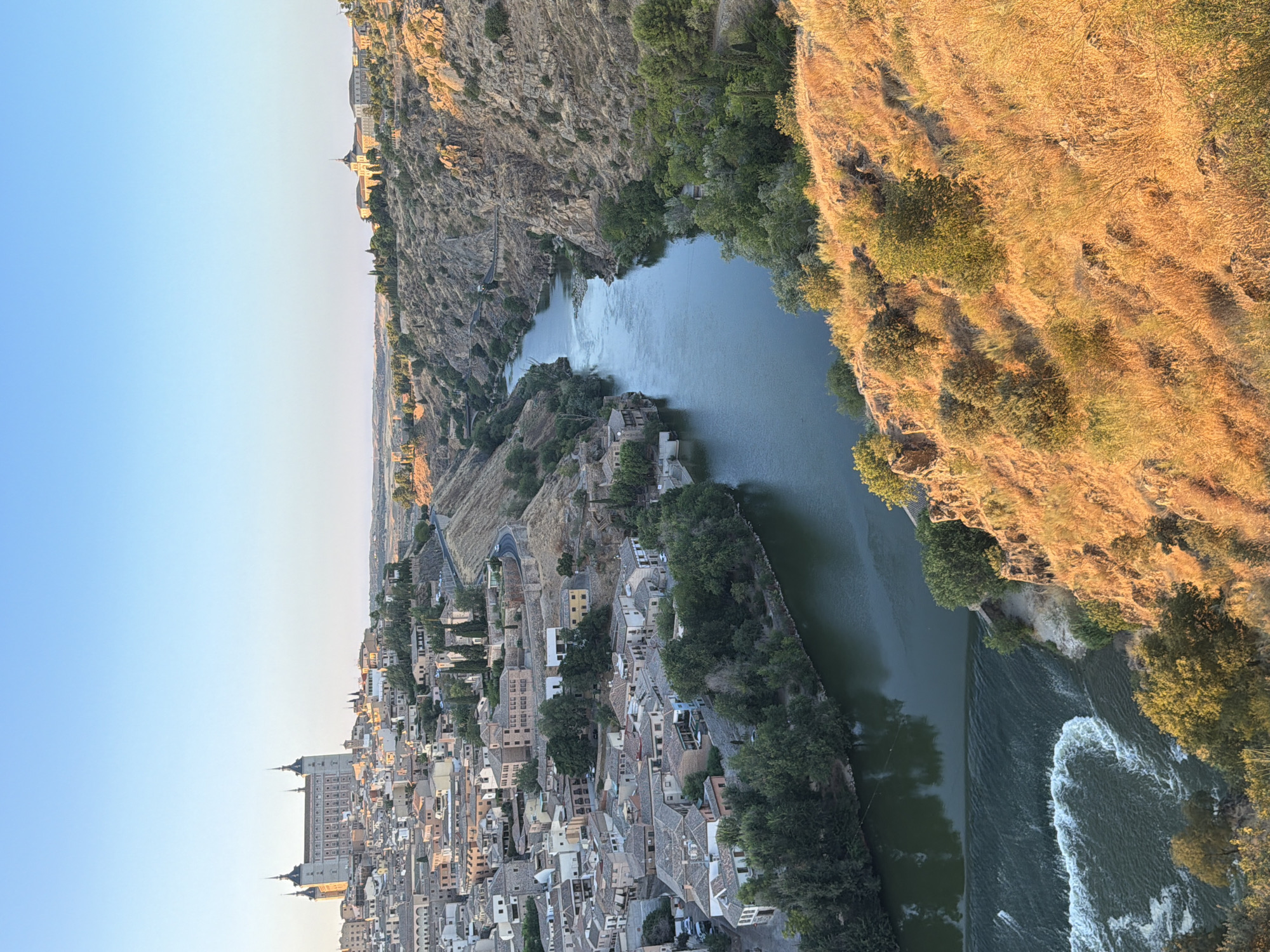This is a high-angle, vertical, full shot of the city of Toledo, Spain, at sunrise. The city is built on a hilltop overlooking the Tagus River, which winds through a deep gorge. The Alcázar of Toledo, a stone fortress with four towers, dominates the skyline on the left. Below the Alcázar, a dense urban landscape of terracotta-roofed buildings stretches across the hillside.
The Tagus River flows from the upper right to the lower left of the frame. The river is dark green and reflects the clear blue sky. On the left side of the river, closer to the foreground, a section of the river forms a small waterfall with white water. Rocky, arid hillsides covered in dry grass and sparse vegetation frame the river on both sides. The sun, low on the horizon, casts a warm, golden light on the right hillside and the city. The sky is a gradient of pale blue, with no visible clouds. No people or animals are discernible in the image.