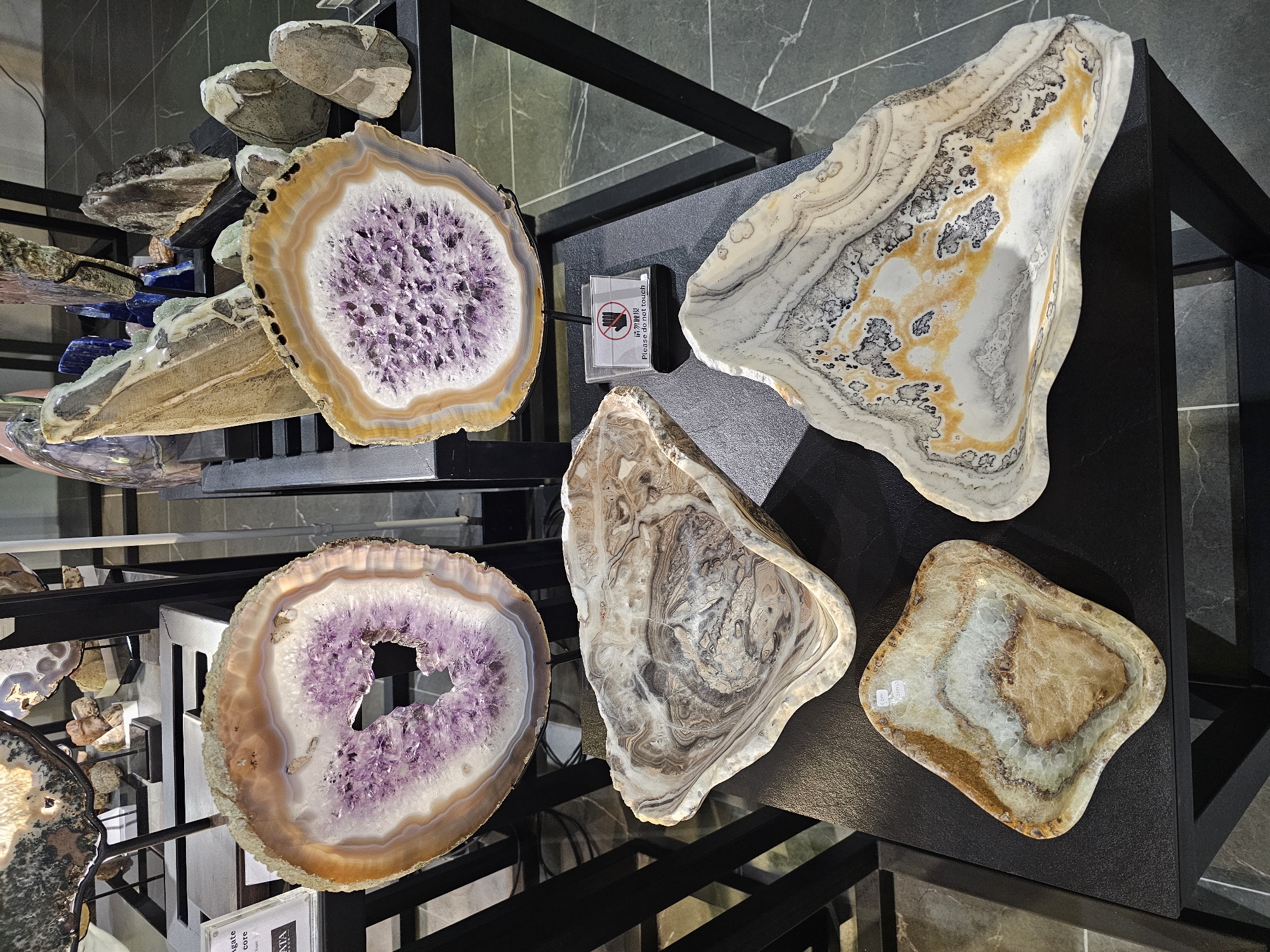 The image displays an assortment of polished agate slices and carved stone bowls arranged on black display stands. In the foreground, a small, irregularly shaped light brown and white stone bowl sits on the lower shelf. To its right, a larger, flat, triangular-shaped stone bowl with swirling patterns of white, grey, and yellow-orange is also on the lower shelf. In the background, two large, circular agate slices are prominently displayed on stands, one with a predominantly white and purple crystalline interior, and the other with a similar purple interior surrounded by bands of white and orange. Several other mineral specimens and carved stone items are visible on shelves in the background, some in focus and others blurred. A small white sign with red text and an icon indicating "Please do not touch" is placed between the agate slices. The scene appears to be an indoor exhibition or retail environment, possibly a museum or a specialized shop. The flooring is dark grey tile. No people are visible in the image. The location is not specified within the image or its provided context.