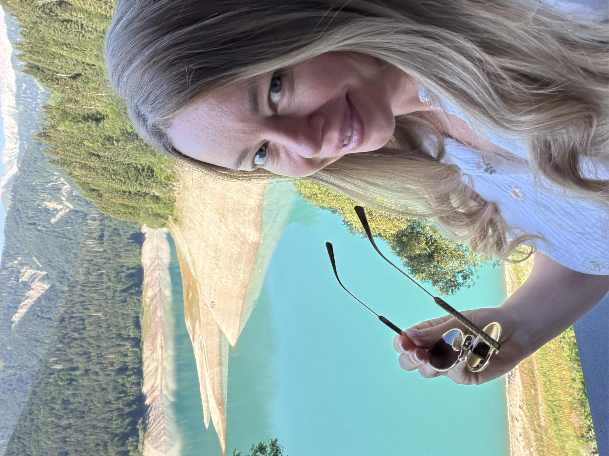 A woman with blonde hair smiles at the camera, holding a pair of sunglasses. She is wearing a white shirt with floral embroidery. The background is a scenic view of a turquoise lake surrounded by lush green forests and mountains. The location is not identifiable as Lateis/Latais, Italy.