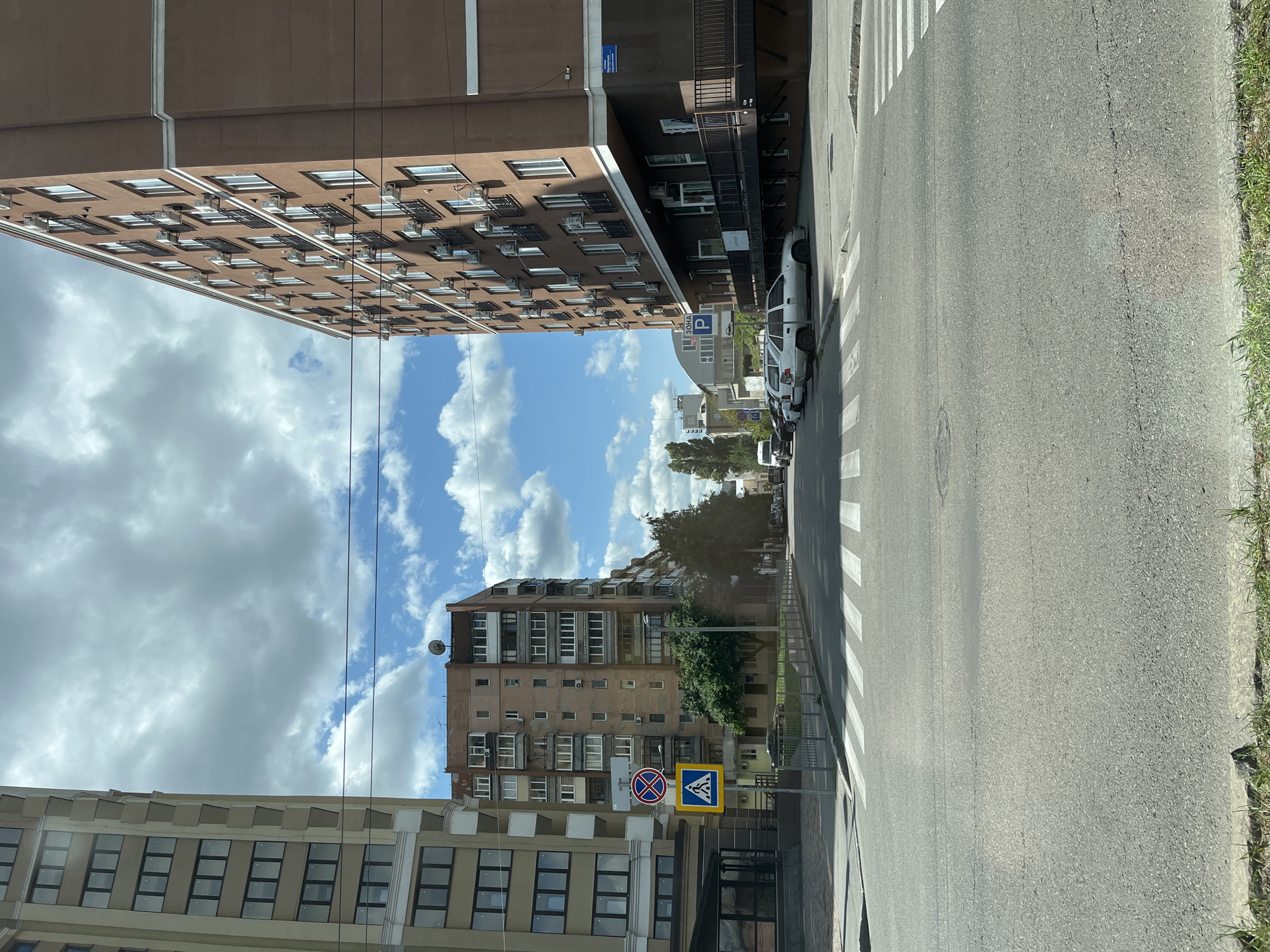 This is a street-level, eye-level, medium-shot, vertical photograph taken on a sunny day in Kharkiv, Ukraine, likely in a residential area. The image captures a crosswalk on a paved road, with two large apartment buildings flanking either side of the street. The building on the left is beige and features a repeating pattern of windows and balconies. The building on the right is a darker, reddish-brown hue with numerous air conditioning units mounted on its facade, and a white decorative trim along its roofline. Several cars are parked along the sidewalk in front of the brown building. A blue and yellow pedestrian crossing sign and a circular red and white "no stopping" sign are visible on the left side of the road, near the pedestrian crosswalk. Overhead power lines are strung across the sky. The sky is partly cloudy, with patches of blue visible between the white clouds.