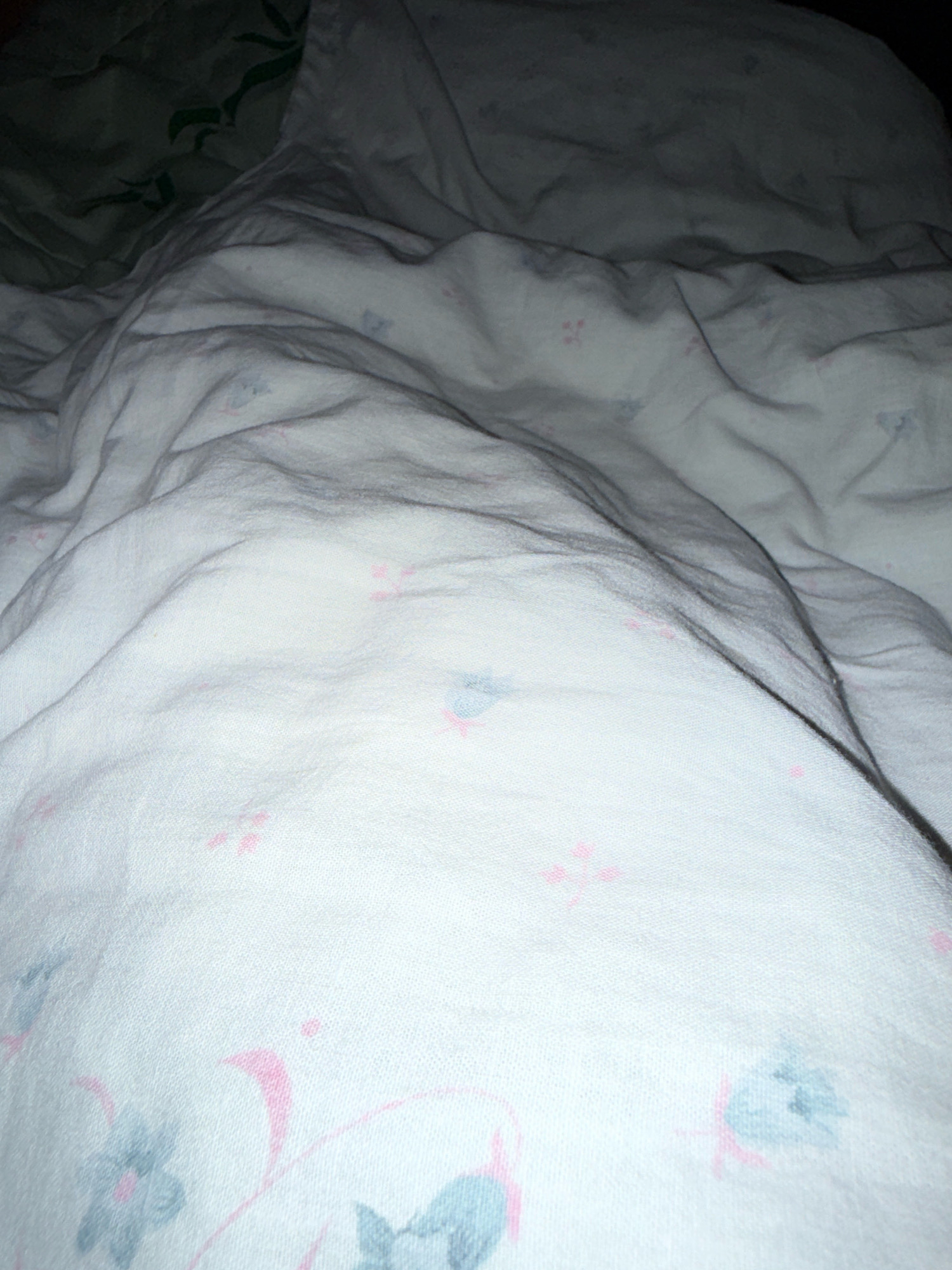 The image shows a close-up view of a rumpled white bedsheet with a subtle floral pattern of pink and blue. The lighting is dim, creating shadows and highlights on the fabric's folds. The environment appears to be indoors, likely a bedroom. There are no people or visible actions captured in the frame. The image is a static depiction of a bedding surface. The location in Zavolzhye, Russia, is not discernible from the visual information presented.