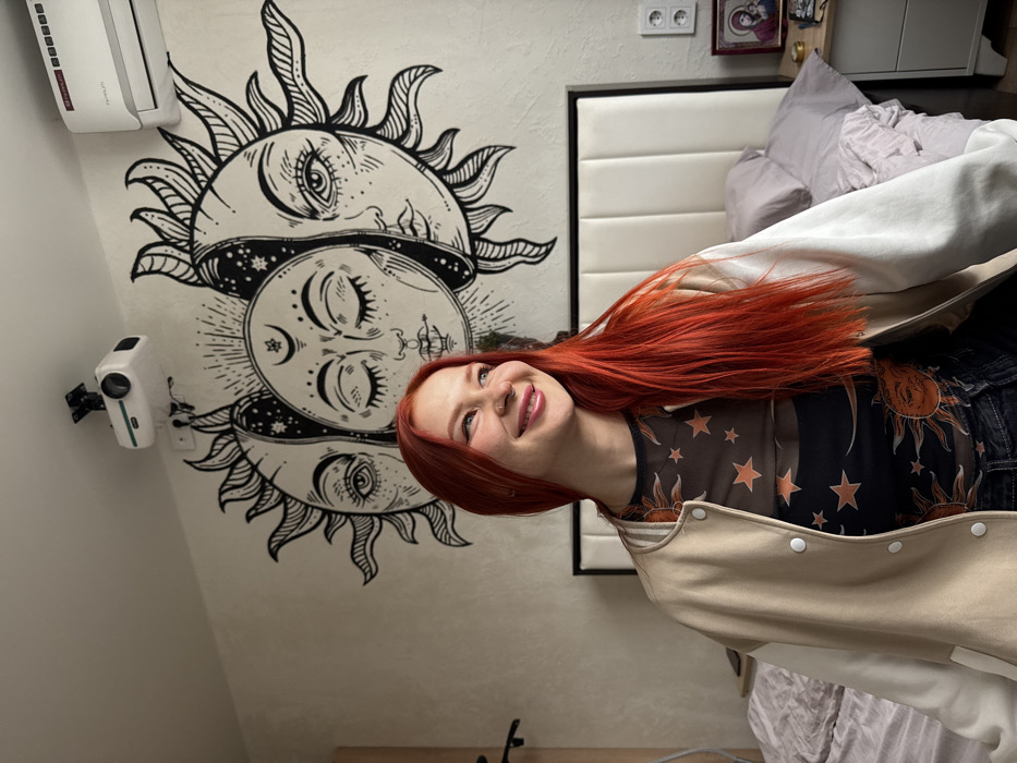 The image depicts a young adult female with long, bright red hair, fair complexion, and a nose piercing, smiling and looking towards the right of the frame. She is wearing a beige and off-white letterman-style jacket over a dark, sheer mesh top patterned with orange celestial bodies including stars and suns. She is seated indoors, specifically within a bedroom located in Rostov-on-Don, Russia.
The background wall features a large, black-and-white mural of three stylized celestial faces: a central moon face with closed eyes, flanked by two sun faces with open eyes. Above the mural, a white projector is mounted on the ceiling to the left, and a white split-system air conditioning unit is mounted on the wall to the right. A bed with a cream-colored, vertically paneled headboard is visible behind the individual, topped with light grey/purple bedding that appears somewhat disheveled. To the right of the bed, a dark nightstand holds a small framed icon, likely depicting the Madonna and Child, and a small cylindrical object. A double electrical outlet is present on the wall above the nightstand. The room's walls are a light, textured neutral color.