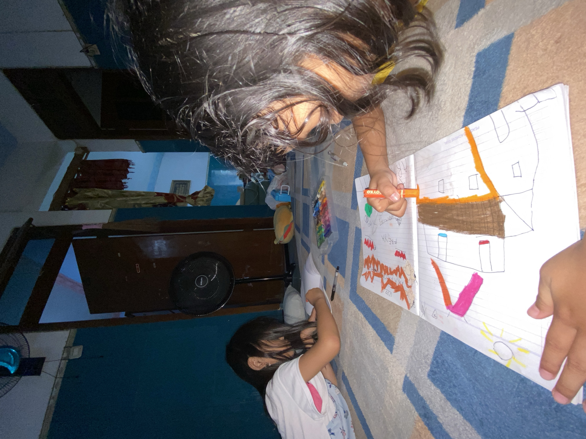 This image depicts an indoor scene in Gelam, Indonesia, featuring two young females engaged in drawing activities on a patterned rug.
The foreground shows a young female lying prone, with dark hair cascading forward. She is wearing a yellow garment, and her right hand holds an orange crayon or marker, actively coloring a drawing of a house with a brown roof and a yellow sun in an open notebook.
To her left, another young female is also lying prone, facing away from the camera. She has dark hair and wears a white short-sleeved top over a pink undershirt, with patterned bottoms. Her left hand holds a pen or marker, drawing in a separate open notebook, which displays drawings including the name "FREYA" and orange scribbles.
Between the two individuals on the rug, a clear plastic container holds a variety of colored markers or crayons. A black marker and a white pen are also on the rug near the background child. A light blue pillow is situated near the background child's head. Further in the background, a yellow stuffed object is visible.
The room features dark teal/blue walls. In the mid-ground, a black Miyako brand pedestal fan stands on the floor. Beyond it, a dark brown wooden door is partially ajar, revealing a brighter blue-lit room beyond. In the upper left background, a wall-mounted oscillating fan with a blue operational light is visible. Through another doorway or opening in the left background, a room with lighter blue walls is partially seen, featuring red and cream-colored draped fabric and a framed object on the wall.