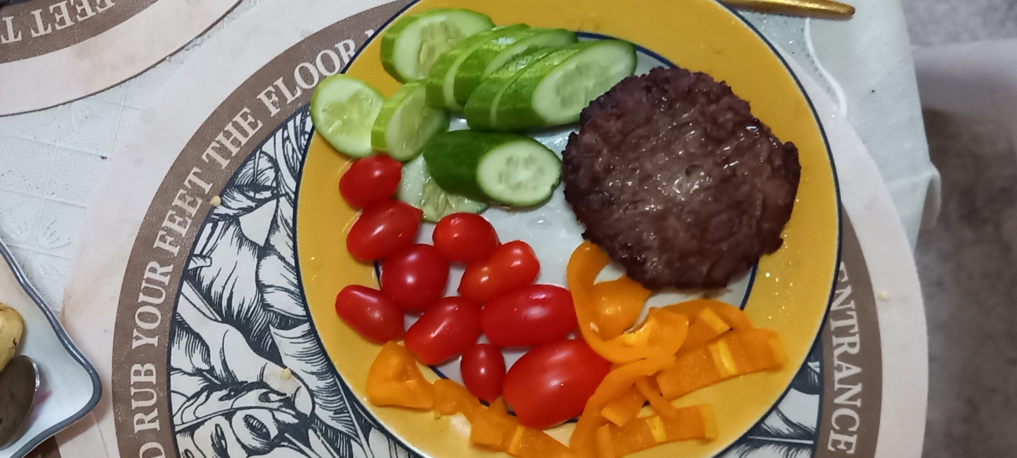 A plate of food is presented on a table in Baan Chaloklum, Thailand. The plate is yellow with a white interior and contains sliced cucumbers, cherry tomatoes, a hamburger patty, and sliced yellow bell peppers. The table has a placemat with black and white illustrations and brown text, which reads in part "RUB YOUR FEET THE FLOOR". The table appears to be indoors, with a white cloth or napkin partially visible.