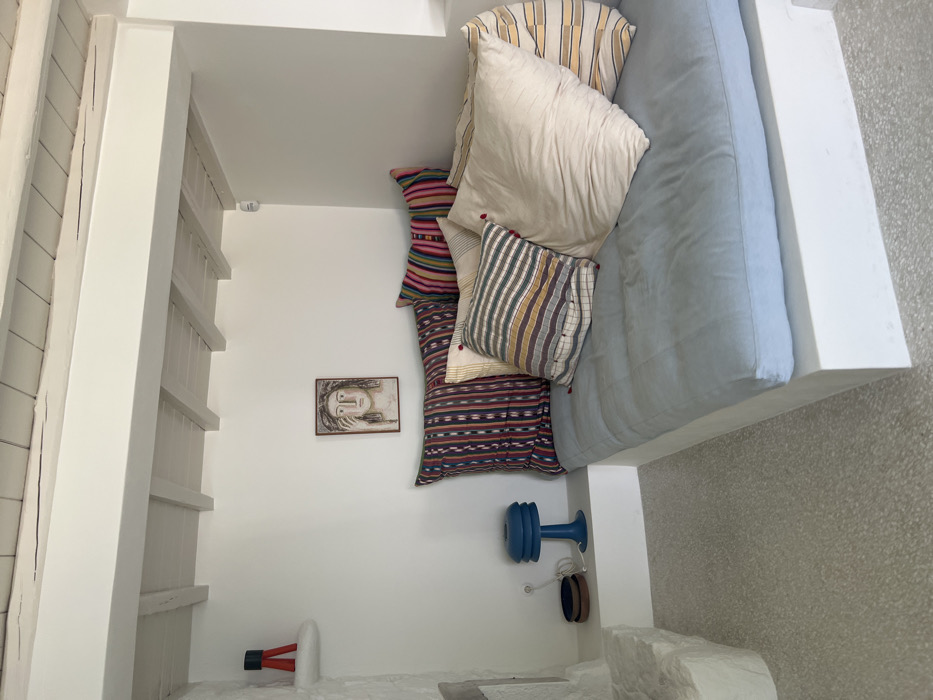 This image displays an interior living space, likely a lounge or seating area, located in Tourlos, Greece. The room features white, smooth plastered walls and a white wooden beamed ceiling, indicative of Cycladic architectural style. The ceiling includes a prominent horizontal beam supporting several perpendicular, smaller white-painted wooden beams. The floor is a light-colored, speckled terrazzo or polished concrete.
A white, built-in L-shaped bench or sofa occupies the right and back walls, furnished with a pale blue, tufted mattress-style cushion. Multiple decorative throw pillows are arranged on the sofa: two large pillows with vertical dark blue, red, and white stripes; one large plain light cream pillow; one medium-sized pillow with horizontal beige, yellow, brown, and grey stripes accented with red tassels; and one large pillow with horizontal yellow and white stripes.
Centered on the back wall above the sofa, a rectangular framed artwork is mounted. It depicts a stylized human face with dark hair, light skin, and one hand raised near the face.
On the left side of the room, a rough-hewn, white stone structure extends from the wall. Above this, a small, white, cantilevered shelf holds a small decorative object consisting of red and dark grey/black geometric shapes. Below, on a white ledge that extends from the built-in sofa, a blue, multi-tiered table lamp is positioned. Next to the lamp, two small, dark, round objects are stacked, and a white electrical cord is visible. A white, cylindrical object, possibly a light fixture or sensor, is affixed to the ceiling in the upper right corner of the room. The scene is illuminated by natural light entering from an unseen source, casting soft shadows. No individuals are present.