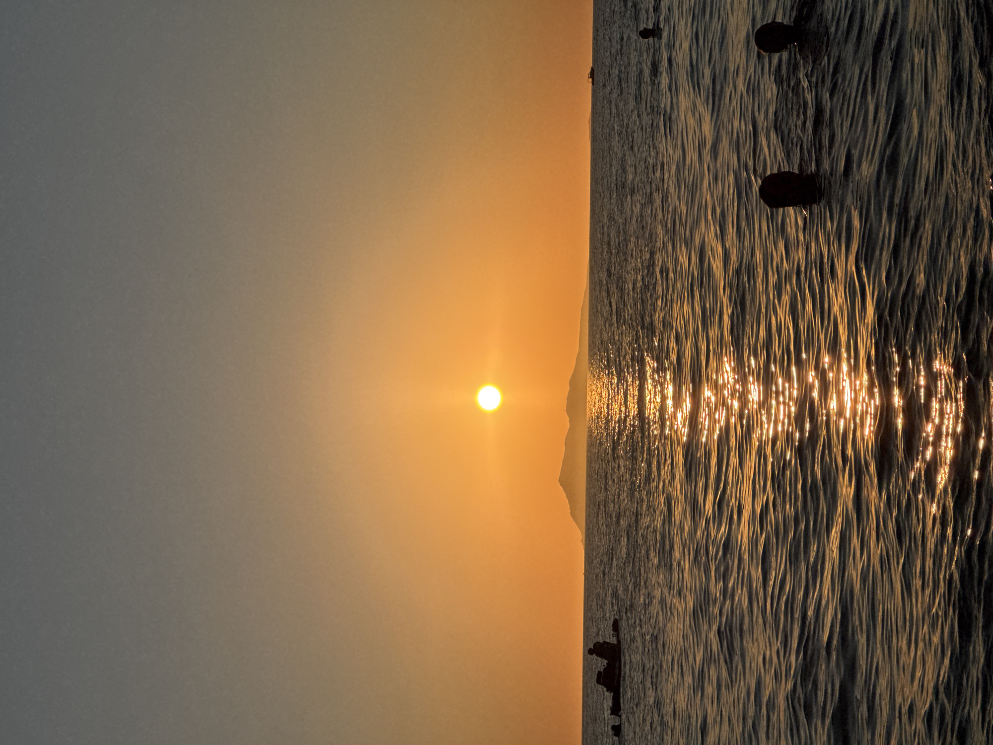 The image depicts a sunset over a body of water. The sun is a bright, glowing orb positioned in the center of the frame, casting a warm orange and yellow hue across the sky and reflecting on the rippling water. In the distance, a silhouetted island or landmass is visible. On the left side of the frame, a small boat with two silhouetted figures is on the water. In the foreground, the heads of several people are visible as they swim or float in the water, silhouetted against the setting sun and its reflection. The water appears dark and somewhat choppy, with streaks of golden light from the sun creating a shimmering effect. The sky transitions from a pale orange near the horizon to a dusky gray at the top of the frame. The scene evokes a sense of peaceful evening relaxation by the sea. There is no visual information in the image that would suggest a location in the city of Urdzhar, Kazakhstan.