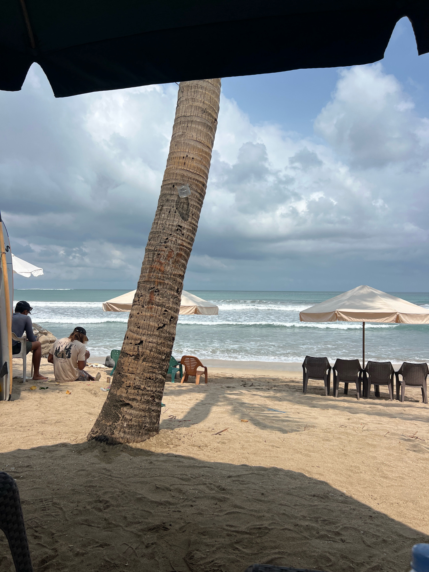 The image depicts a beachfront scene in Kuta, Indonesia, under a cloudy sky. In the foreground, a large palm tree trunk dominates the frame, with its shadow stretching across the sandy beach. To the left, two individuals are seated on chairs, facing away from the camera, with a surfboard leaning against a structure. In the middle ground, several beach chairs are arranged under a beige umbrella, with the ocean waves gently rolling onto the shore in the background. Additional chairs and a second umbrella are visible further down the beach. The overall atmosphere suggests a relaxed, tropical setting.