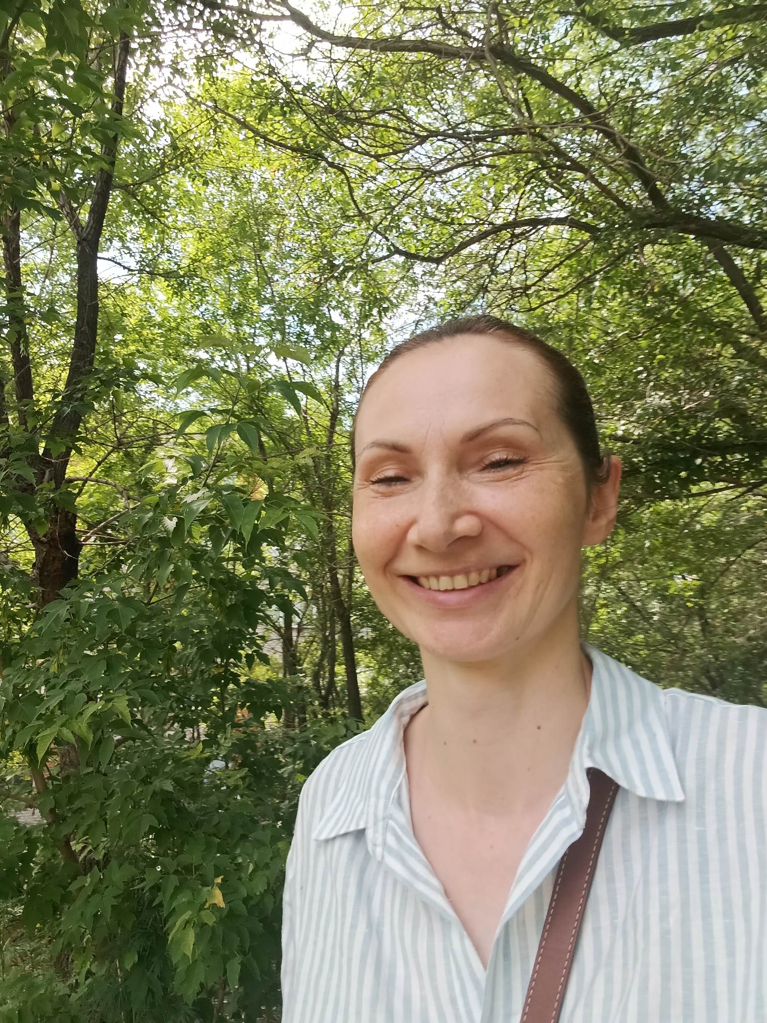 A woman with fair skin, brown hair pulled back, and freckles smiles at the camera. She is wearing a light blue and white striped collared shirt, and a brown strap is visible across her shoulder. The background is filled with lush green trees and foliage, suggesting an outdoor setting. The lighting indicates it is daytime, with sunlight filtering through the leaves. The scene is likely a park or a wooded area within the city of Komsomolsk-on-Amur, Russia, given the presence of mature trees and the commonality of such natural spaces within urban environments. There are no other people or significant objects in the frame.
