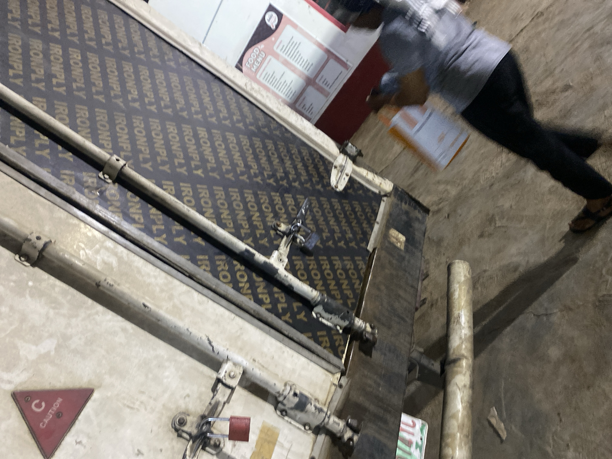 The image depicts an outdoor or semi-enclosed commercial area within the city of Ikorodu, Nigeria, under artificial lighting, indicated by prominent shadows.
The scene features a light-colored, heavily worn, and soiled metallic or painted surface, possibly part of a vehicle, container, or industrial door, positioned on the left side. Attached to its upper portion is a red triangular caution sign with a white "C" and "CAUTION" text. Below the sign, a heavy-duty metallic latching mechanism is secured with a red padlock.
Adjacent to this, a large, dark-colored plywood panel, extensively branded with repeating white "IRONPLY" text, forms a vertical barrier or wall. Another metallic latching mechanism, secured by a darker padlock, is visible on this plywood surface. Several metallic pipes or bars run vertically in the mid-ground, and one horizontal pipe lies on the dark, uneven concrete or asphalt ground.
To the right, a "FOOD MENU" board, colored pink and white, is affixed to a red vertical surface, indicating a food service establishment. In the lower right, a single individual, blurred by motion, is observed walking away from the camera. The person is wearing a light-colored short-sleeved top, dark trousers, and sandals, and is carrying a light-colored, rectangular object, possibly a package or tray.