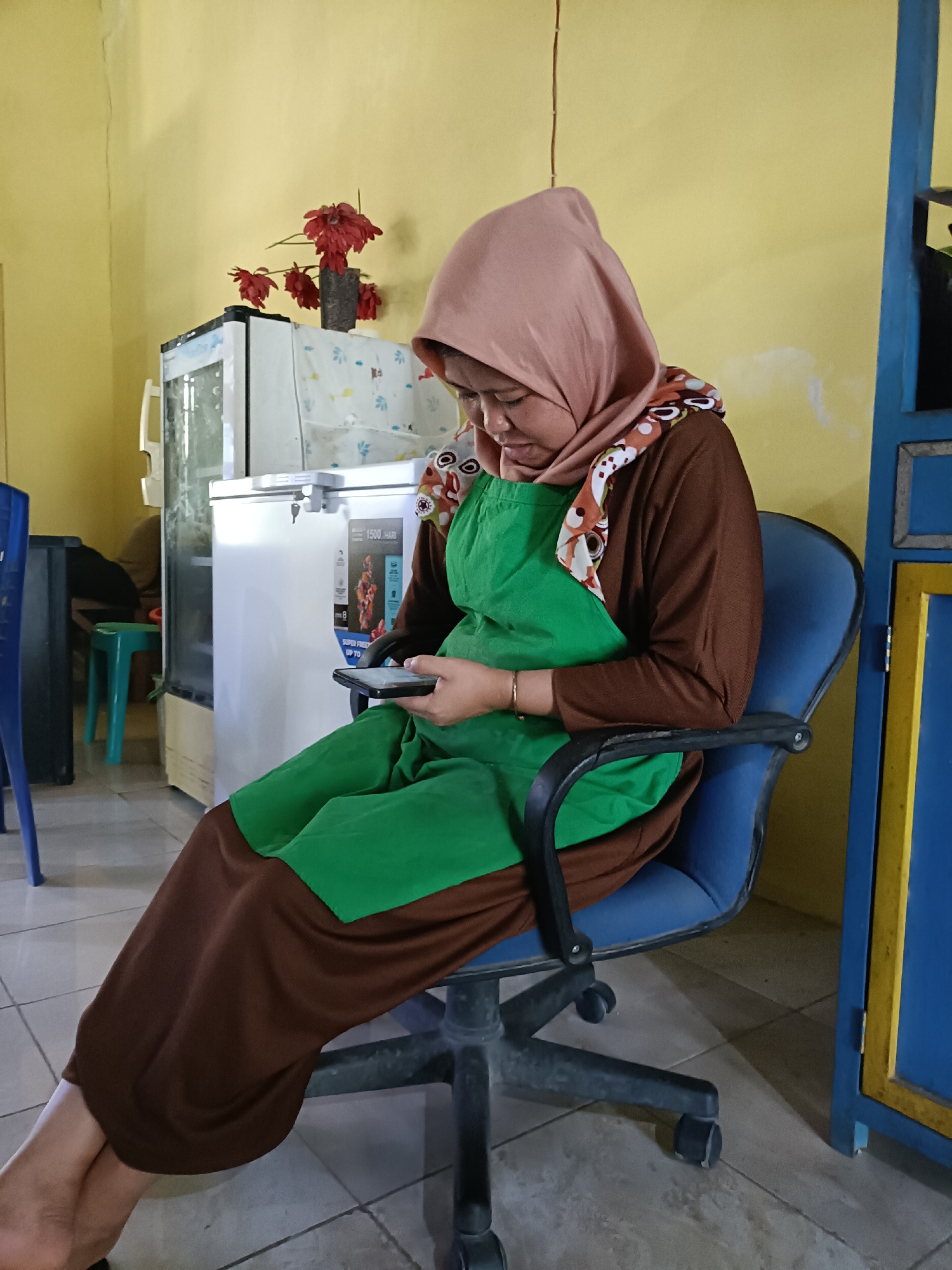 A woman wearing a pink hijab and a brown long-sleeved shirt with a green apron is sitting on a blue office chair in what appears to be a room in Pordapor, Indonesia. She is looking down at her phone, which she holds in her hands. Her legs are crossed. In the background, a white refrigerator is visible, and to the right, a blue cabinet with yellow trim is partially in view. The walls are painted yellow. A vase with red flowers sits on top of the refrigerator.