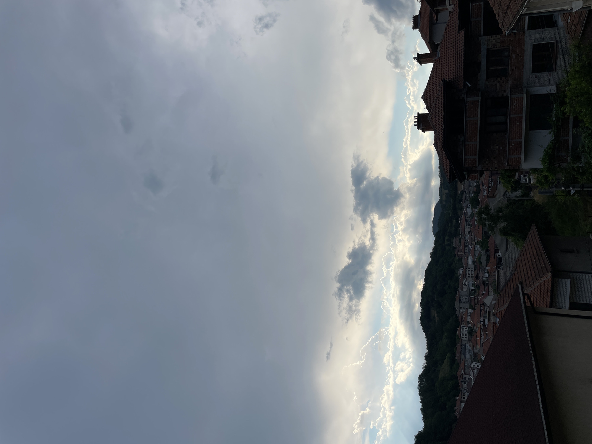 The image depicts a view of the town of Zlatograd, Bulgaria, from an elevated perspective. The foreground is dominated by the angled rooftops of buildings, some appearing to be under construction due to exposed brickwork. Beyond the immediate rooftops, a densely packed town unfolds, with numerous buildings featuring terracotta tiled roofs, nestled on a hillside. The hillside is covered in lush green trees, creating a stark contrast with the man-made structures.
The sky takes up a significant portion of the frame and is filled with dramatic clouds. The left side of the sky is overcast with a dense, grayish layer of clouds, suggesting a subdued or impending weather event. In contrast, the right side of the sky is brighter, with sunlight illuminating the edges of fluffy white clouds, creating a striking halo effect. This suggests a dynamic weather pattern, possibly transitioning from overcast conditions to a clearer, sunnier sky.
The time of day appears to be late afternoon or early evening, given the angle of the light and the nature of the cloud formations. The overall mood is one of serene observation, capturing a moment of natural beauty against the backdrop of a charming European town. No people are visible in the image, and there is no visible text.