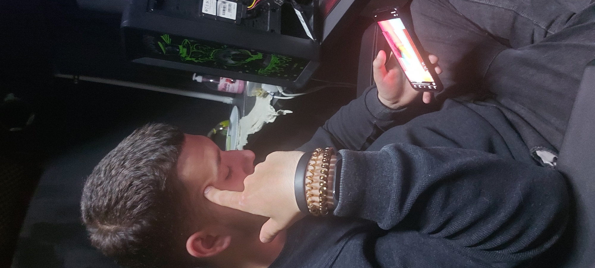 A man is seated indoors, viewing a smartphone. He is wearing a black long-sleeved shirt and dark jeans. On his left wrist are several bracelets, including a beaded one and a dark band. His right hand is touching his forehead near his eye. To the left of the man is a computer tower case with a green dragon design. A white bottle and other miscellaneous items are visible on the surface next to the computer. The scene appears dimly lit. It is not possible to determine the location within Tashkent, Uzbekistan from the given image.