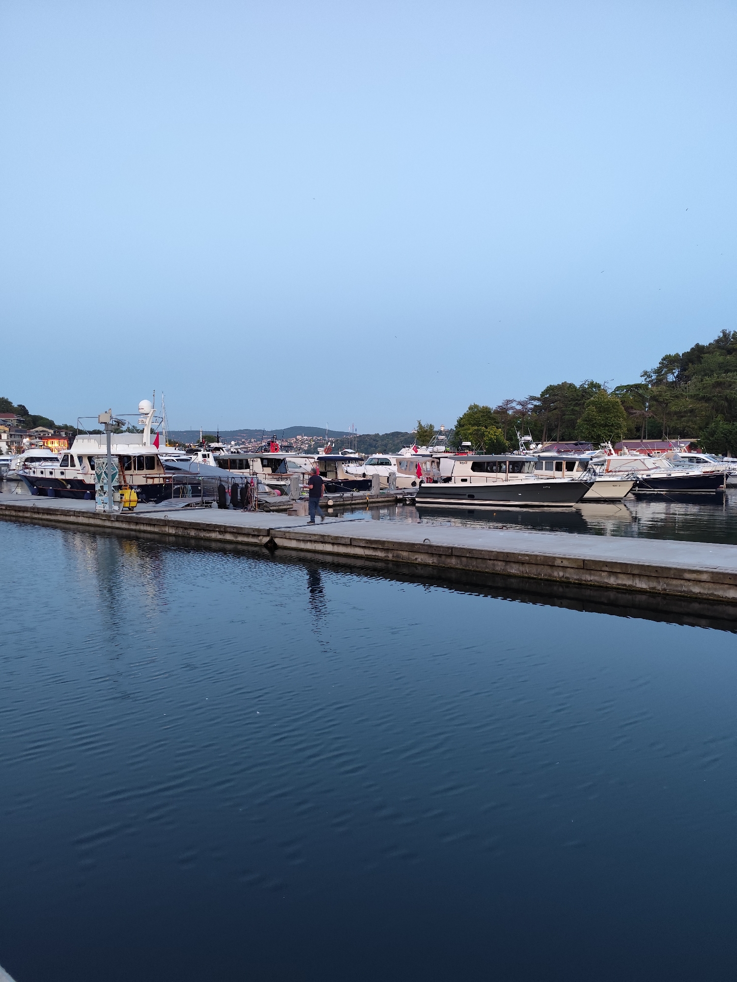 The image depicts a serene harbor scene in Sarıyer, Turkey, likely during twilight. A long concrete pier extends into dark blue water, its surface rippled by a gentle breeze. Several yachts and boats are moored alongside the pier, their structures varying in size and design. One prominent boat, light blue and white, has a red flag flying from its mast. A lone individual, dressed in dark clothing, walks along the pier, casting a faint reflection on the water below. Beyond the boats, a lush green hillside is visible, dotted with trees and some distant buildings, suggesting the harbor is situated in a coastal town. The sky above is a pale, clear blue, transitioning into a deeper hue at the horizon.