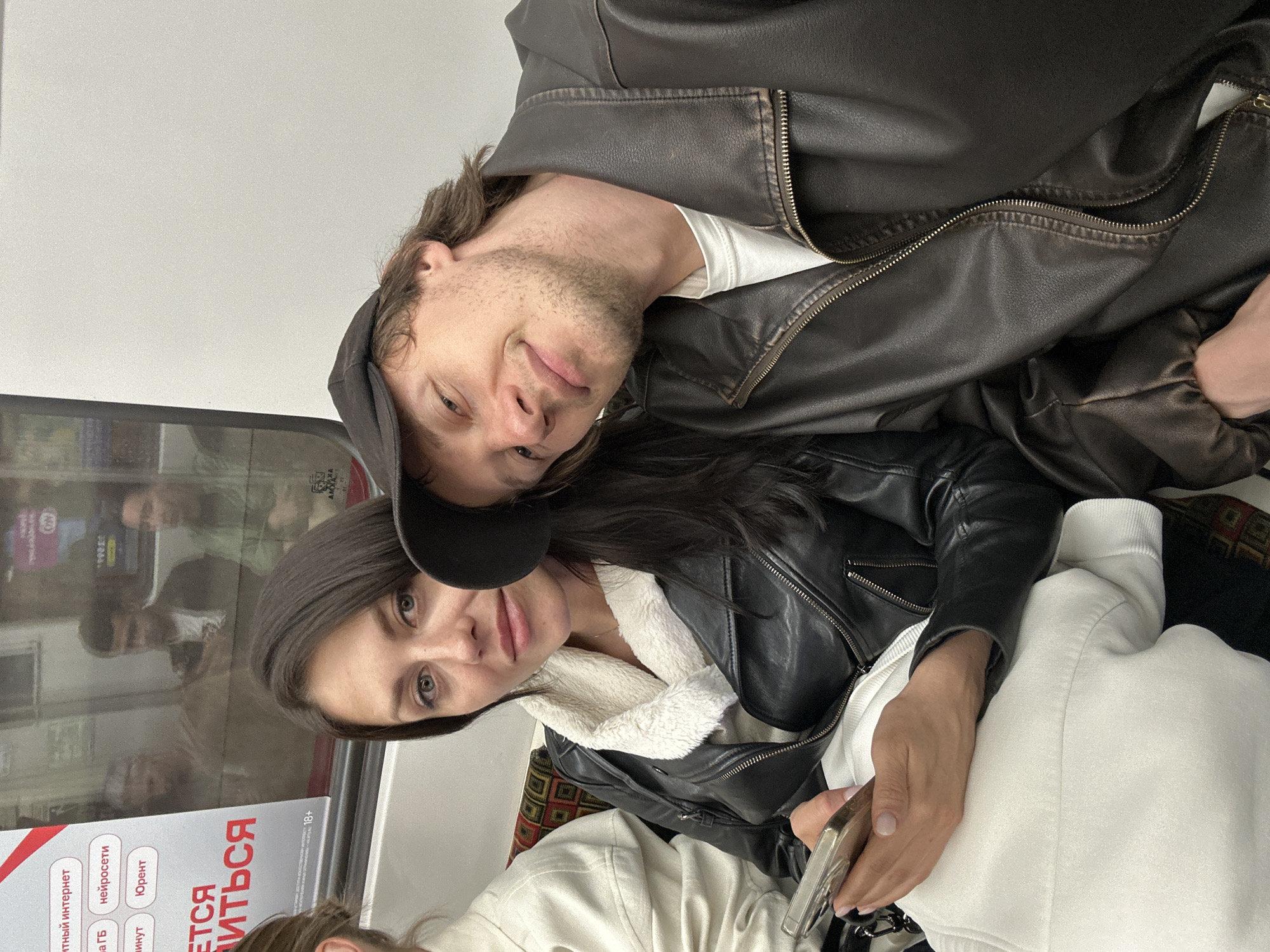 Two individuals, a woman and a man, are seated next to each other in what appears to be a subway car. The woman, in the foreground on the left, has dark hair and is wearing a black leather jacket with a white fuzzy collar. She is looking at her phone, which she holds in her left hand. The man, on the right, is wearing a dark baseball cap and a brown leather jacket. He has his head resting on the woman's shoulder and is looking towards the camera with a slight smile. In the background, reflected in the window of the subway car, several other passengers are visible. The interior of the subway car shows seating with patterned upholstery and a white wall. On the left side of the image, there is a window with text and graphics, some of which are in Russian. The overall scene suggests a casual journey on public transportation.