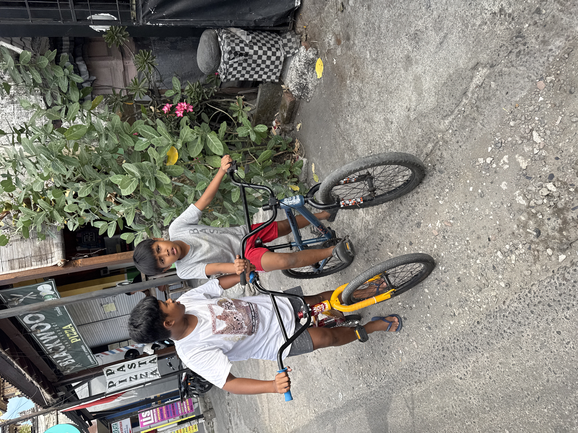The image captures two young boys on bicycles in front of a commercial establishment with "BLACKWOOD PIZZA" signage in Denpasar, Indonesia. The boy on the left, wearing a white t-shirt and grey shorts, is on a yellow and red BMX bike. The boy on the right, in a grey t-shirt and red shorts, is on a blue BMX bike. Both boys appear to be looking towards the camera. The environment includes a paved street with visible cracks, a building with shuttered windows, and lush green foliage with pink flowers. A checkered cloth covers a structure to the right of the children, and a large, dark stone sits atop it. Signs for "PASTA" and various products are visible on the building to the left. A motorcycle is parked in the background.
