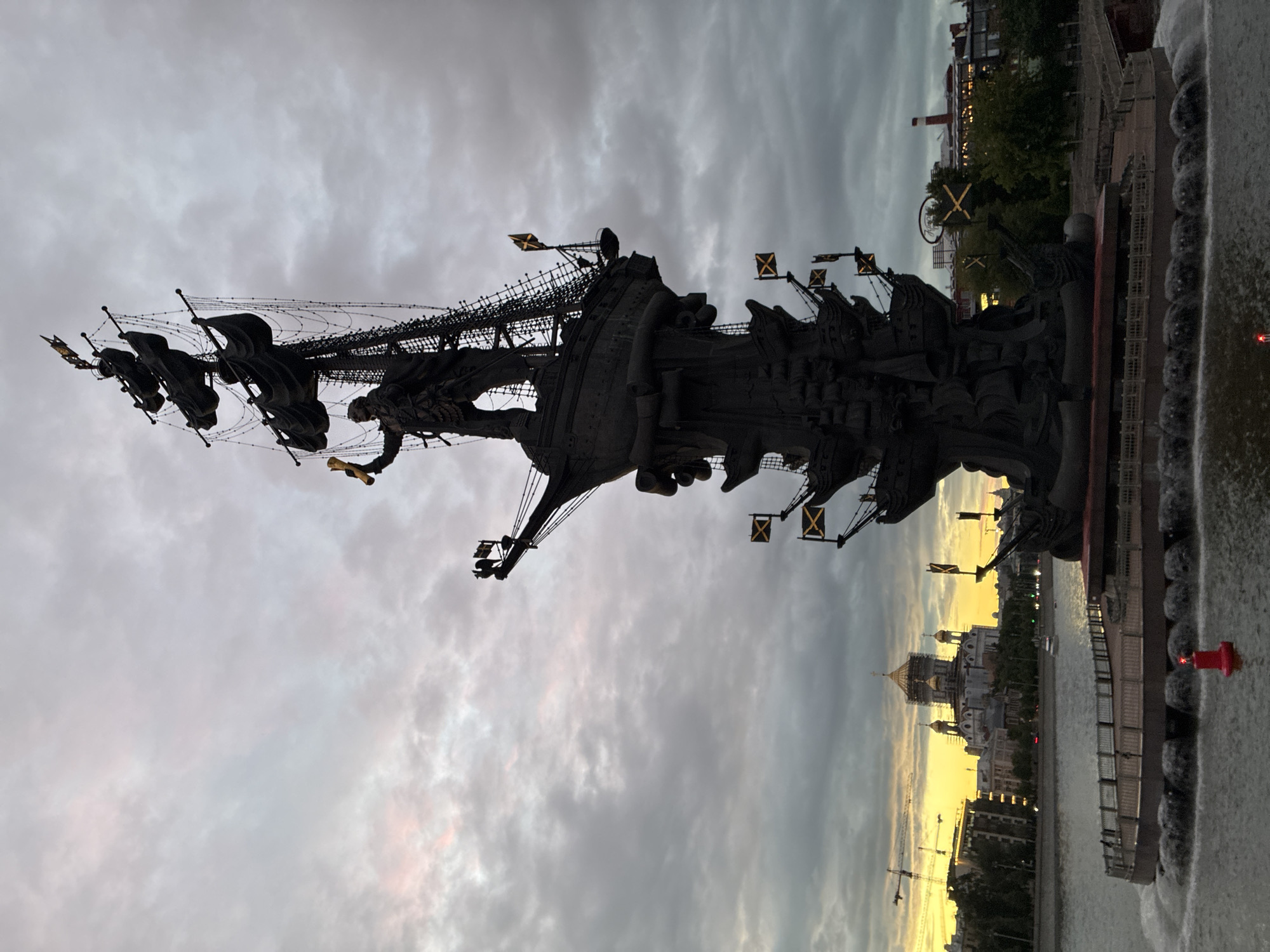 Here is a description of the image:
The image depicts the Peter the Great Statue in Moscow, Russia, at dusk or dawn, against a cloudy sky with a hint of yellow light near the horizon. The statue, a large bronze monument of a ship with figures, stands in the foreground with the Moscow River on the right. The Cathedral of Christ the Savior is visible in the background, along with other buildings and construction cranes. The image appears to be taken at a low angle, looking up at the statue. The water has some surface agitation. The edge of a platform can be seen along the water line to the right. A single red marker is at the right edge of the frame.