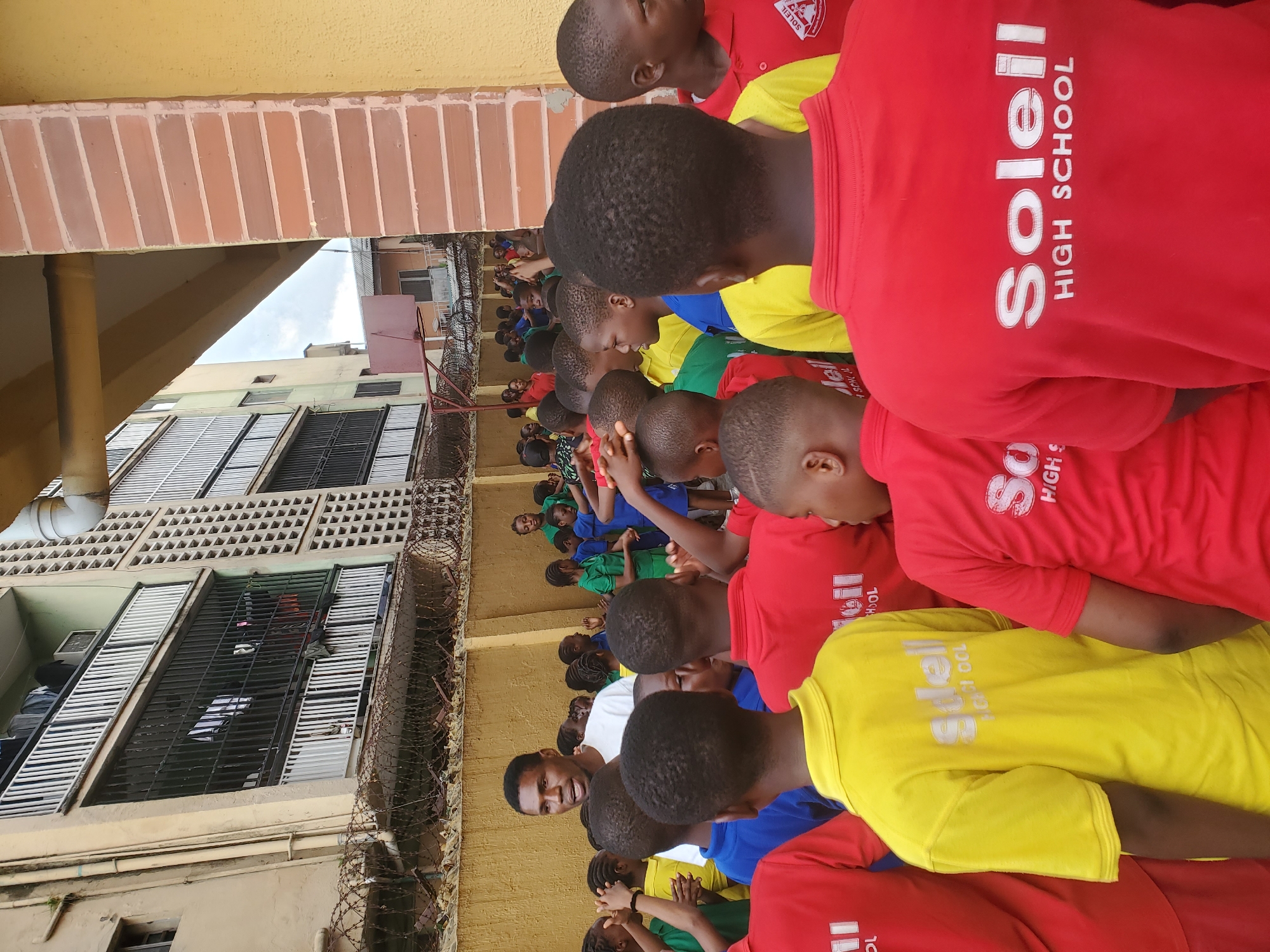 The image depicts a group of children, likely students of "Soleil High School," gathered outdoors in Agani, Nigeria. Most of the children are wearing red, yellow, blue, and green shirts, seemingly school uniforms. A man wearing a white shirt stands amongst the children. Buildings, one of which has windows with security bars, are visible in the background, along with barbed wire along a wall.