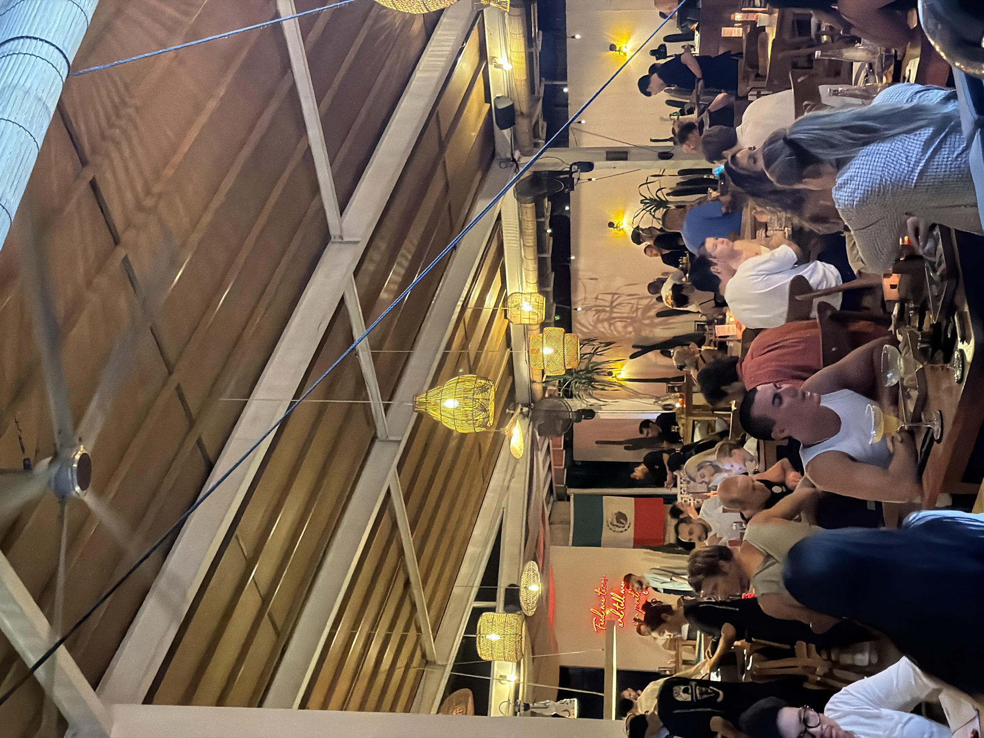 The image depicts the interior of a busy restaurant in North Kuta, Indonesia, with a vibrant atmosphere and a significant number of patrons. The ceiling features a modern design with wooden slats and exposed beams, illuminated by several woven pendant lights that cast a warm glow. A large ceiling fan is visible, indicating a need for air circulation in the tropical climate.
The restaurant appears to be decorated with a Mexican theme, as evidenced by the Mexican flag prominently displayed on the wall and neon signage that reads "Te amo tacos" and "and tell me..." suggesting a casual and lively dining experience. Several potted plants, including cacti, are placed around the establishment, adding to the ambiance.
The foreground and midground are populated with diners seated at wooden tables. People are engaged in conversation, eating, and drinking. The individuals are diverse in age and gender, dressed in casual attire suitable for a restaurant setting. In the foreground, a person with glasses is visible, looking towards the left, while another person with dark hair is seated to their right. Further into the scene, a man in a white tank top is speaking, with a drink in front of him. Other groups of people are seen conversing and enjoying their meals at various tables. The overall scene captures a lively evening at a popular dining spot in North Kuta.