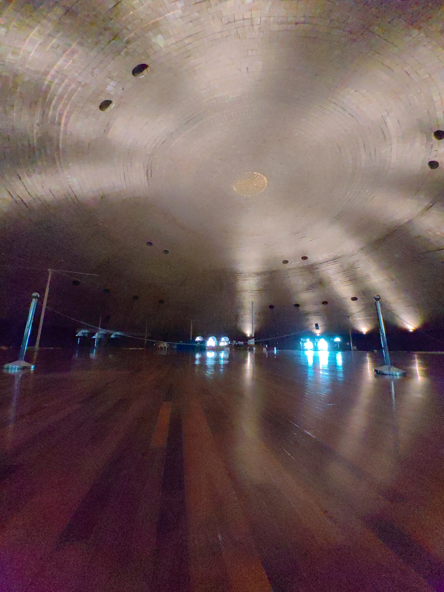 The image depicts the interior of the Great Hall at the Chhatrapati Shivaji Maharaj Vastu Sangrahalaya (CSMVS) museum in Mumbai, India. The camera is positioned low, looking upwards towards the domed ceiling. The ceiling is made of curved, rectangular tiles and features several circular openings. In the center of the ceiling, there is a circular decorative element with radiating lines, possibly a light fixture or ornamentation.
The floor is made of polished wood, reflecting the light from the ceiling and the distant lights. Several vertical stanchions with thin wires strung between them are visible, creating a sense of division or guidance within the space. In the distance, along the curved wall, are arched openings, some of which are illuminated with bright, cool-toned lights, creating vibrant reflections on the floor. The overall atmosphere is one of spaciousness and architectural grandeur, with dramatic lighting casting long shadows. There are no people clearly visible in the image.