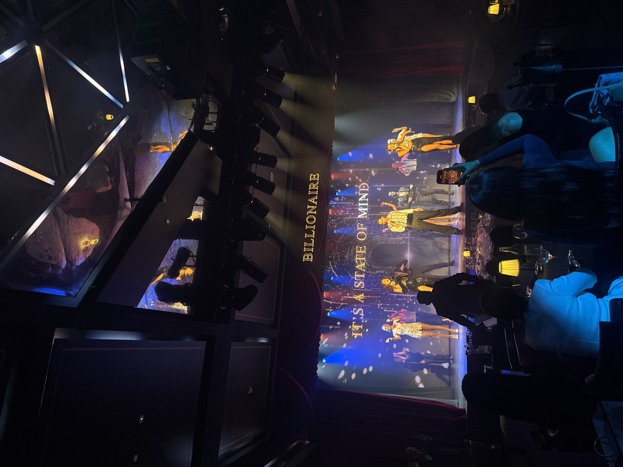 The image captures a performance titled "Billionaire" at a venue likely located in Dubai, United Arab Emirates. The scene depicts a stage production with performers dressed in glamorous costumes, illuminated by stage lighting against a backdrop of visual effects. Audience members are seated in the foreground, some using mobile phones to capture the performance. The venue appears to have a tiered seating arrangement, and the lighting fixtures above the stage are visible. The overall atmosphere suggests an upscale entertainment setting.