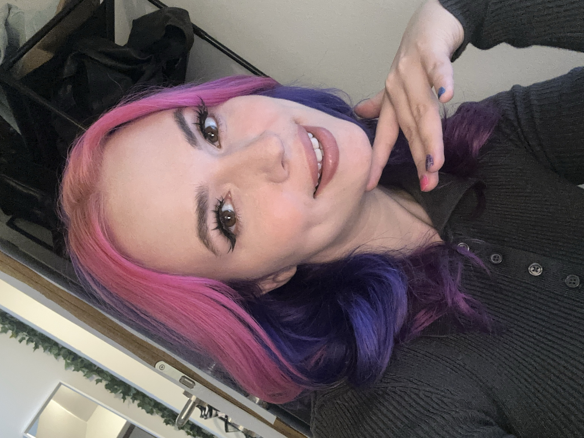 A woman is facing the camera. Her hair is dyed pink and purple. She is wearing a dark grey button-up shirt. She is indoors. The image is rotated. The city is not visible in this image.