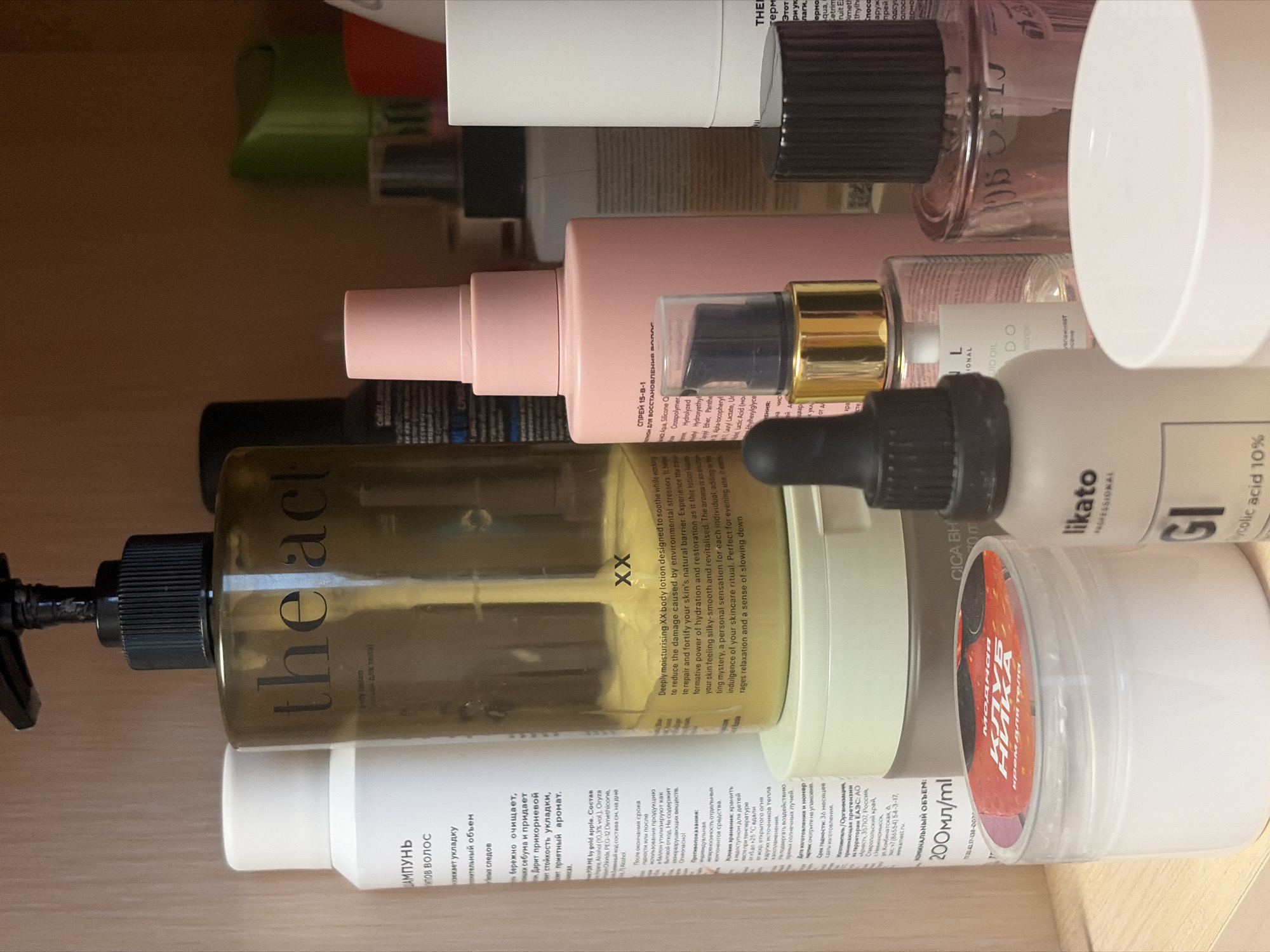 The image shows a close-up of various cosmetic and personal care products, seemingly stored together in a drawer or cabinet. Brands include "the act," "likato PROFESSIONAL," "GI glycolic acid 10%," "Moricia," and some with Russian text. The "the act" product is described as a body lotion, while another bottle with Russian text appears to be a shampoo. The products are stacked close together, suggesting they are in a storage space. The label on "the act" product indicates its purpose is to soothe skin and reduce damage from environmental stressors. Some labels are in Russian. The image was taken in Chaikovsky, Russia.