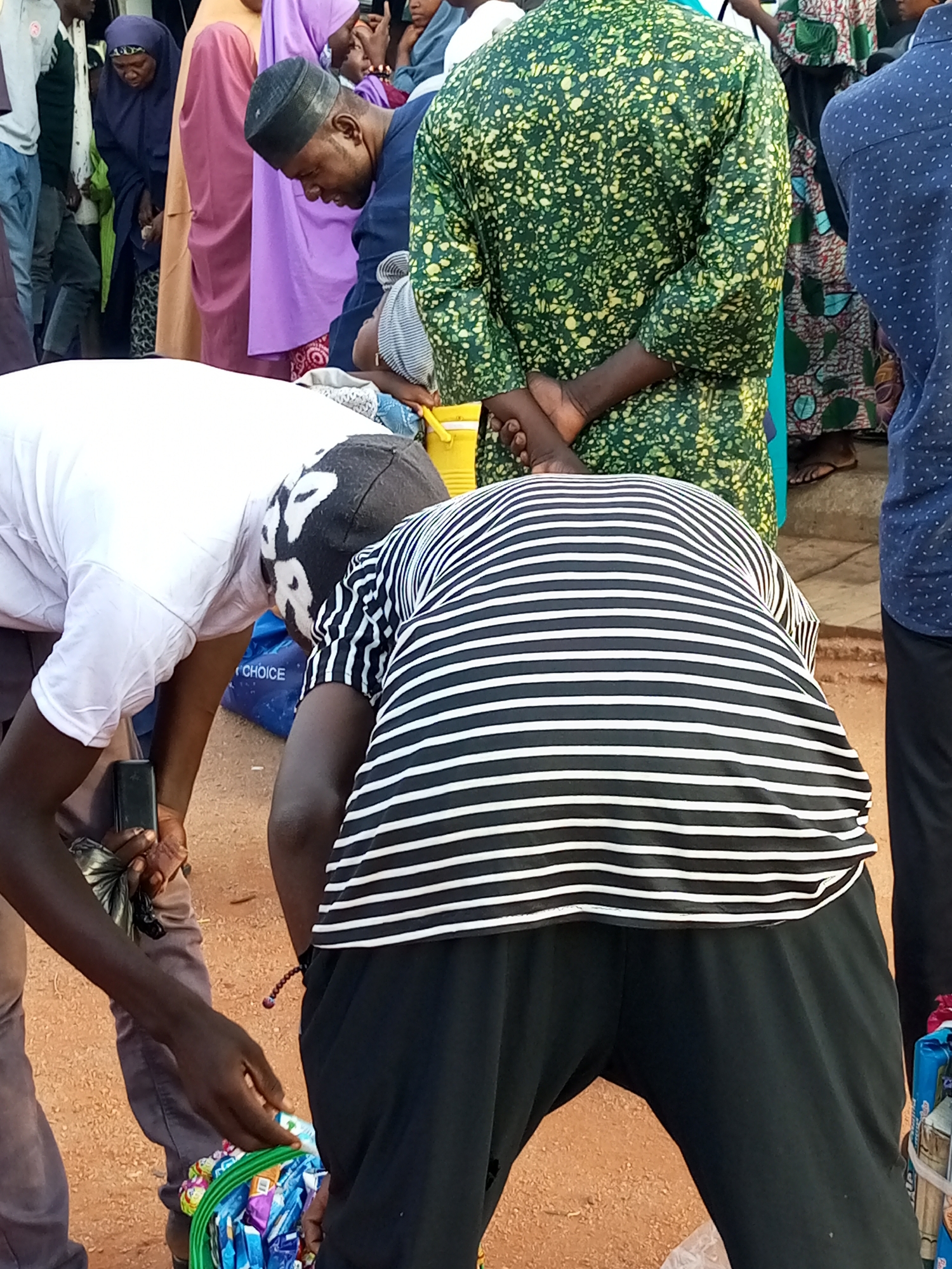 This image captures a scene in the city of Gombe, Nigeria, likely on an outdoor market day. The focal point is a group of people gathered closely together. In the foreground, two individuals are bent over, one wearing a white t-shirt and the other a black and white striped t-shirt. The person in the striped shirt is reaching towards a green basket filled with various packaged items, possibly snacks or small goods. To the left, another person in dark pants and a white t-shirt is also leaning forward, holding a black object in their left hand, possibly a phone.
Behind them, several other individuals are visible. One man wears a dark blue cap and a navy blue robe, looking downwards. Another person is in a vibrant green patterned kaftan with yellow speckles. Further back, a woman in a purple hijab and a pink shawl is seen, alongside other individuals in traditional attire. The ground appears to be dirt or sand, typical of outdoor markets. The overall atmosphere suggests a moment of transaction or focused activity within a community gathering.