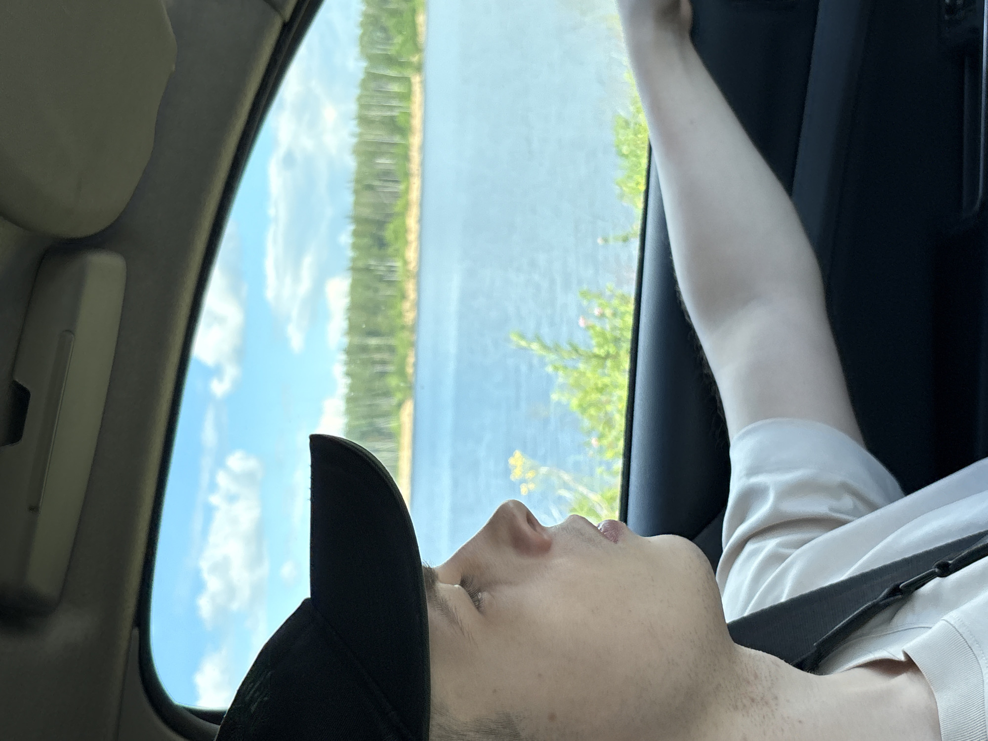 A young Caucasian male, wearing a black baseball cap and a light-colored t-shirt, is seated in the passenger seat of a vehicle, looking out the side window. His right arm rests on the open window frame. Outside the car, a body of water is visible, with a shoreline lined with trees and a cloudy sky overhead. The scene appears to be a scenic route, possibly on the outskirts of Manushkino, Russia, given the natural landscape. The overall impression is one of a leisurely drive through a picturesque environment.