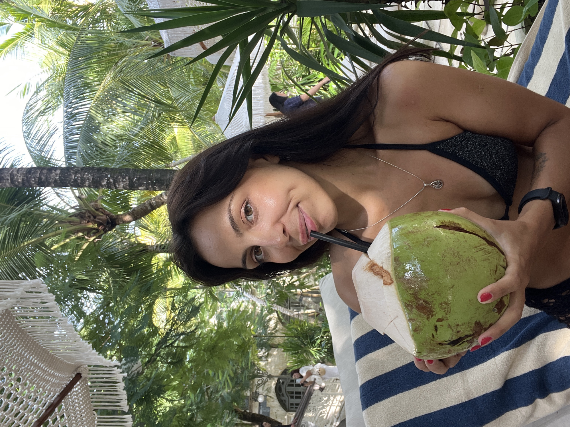 A young woman is captured in a close-up, diagonal shot, relaxing outdoors in a tropical setting, likely Kuta Selatan, Indonesia. She is looking directly at the camera with a playful smile, her tongue subtly touching the black straw inserted into a green coconut she holds. Her long, dark hair frames her face, and she wears a black, sparkly bikini top paired with a silver pendant necklace. Red nail polish adorns her fingers as she grips the coconut, and a black smartwatch is visible on her left wrist.
She appears to be reclining on a blue and white striped towel or fabric. The background is filled with vibrant green tropical foliage, dominated by large palm trees with distinct fronds and trunks, suggesting a bright and sunny day. To the left, a portion of a white macrame or woven structure, possibly a hammock or decorative chair, is visible. In the softer focus background, hints of other people and architectural elements, possibly part of a resort or outdoor leisure establishment, can be discerned, including a round decorative feature. The scene conveys a relaxed, vacation-like atmosphere. There is no visible text in the image.