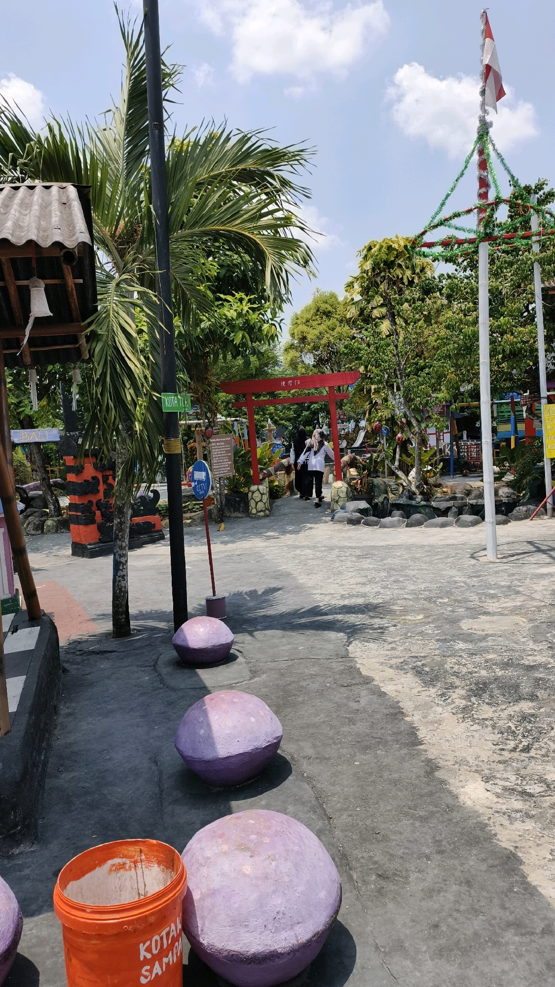 This is a medium-angle, eye-level, full shot of a paved outdoor area with several decorative elements and a few people. In the foreground, a bright orange bucket labeled "KOTAK SAMPA" is on the left, next to three large, lavender-colored, spherical objects placed in a line on the ground. The ground is a mixture of gray pavement and textured concrete.
To the left, there is a partial view of a building with a grey, corrugated roof and a white bell hanging from it. A dark pole, possibly a lamppost, stands behind the bucket and the spherical objects. Further back, a tall palm tree with green fronds is visible, its trunk partially obscured by the pole. Behind the palm tree, a red torii gate stands, with two people in dark clothing walking through it. One person is wearing a white shirt.
To the right of the torii gate, there are trees and foliage. A flagpole with an Indonesian flag is visible in the upper right corner, adorned with festive green and red tinsel. The sky is blue with scattered white clouds. Signs are present in the scene, including a green sign with the words "KOTA TU..." on the pole and a blue circular sign with a CCTV symbol. The overall environment appears to be a public space or park, possibly in the city of Air Paoh, Indonesia.