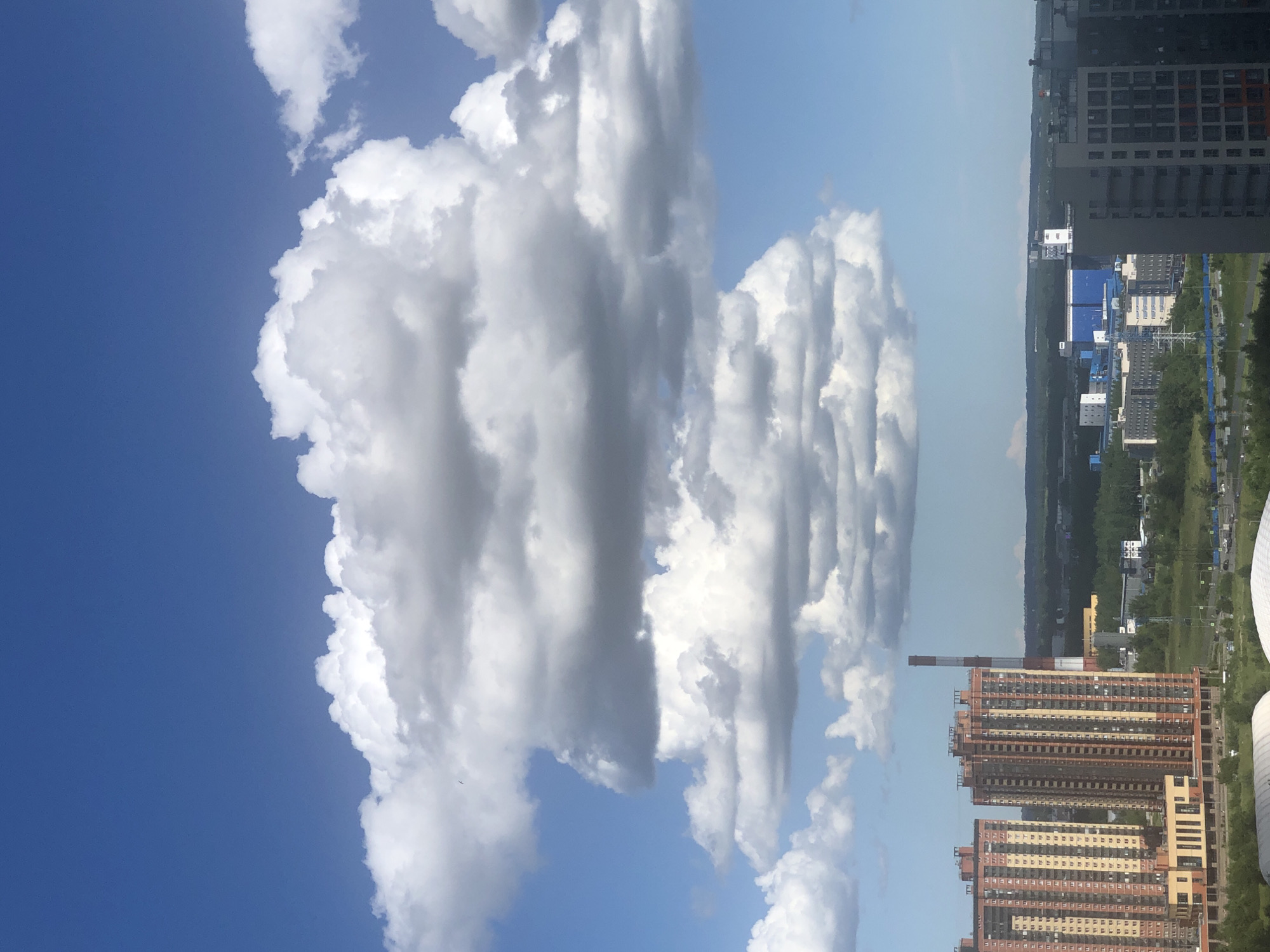 A high-angle, long shot captures a cityscape under a vast, clear blue sky dotted with large, fluffy cumulus clouds. In the foreground, a series of tall residential buildings are visible, characterized by their brick exteriors in shades of orange and beige, featuring numerous windows. Adjacent to these buildings is a tall, red and white striped industrial smokestack. Beyond the residential towers, a sprawling landscape of green trees and industrial structures unfolds. Further in the distance, more buildings and a highway can be seen, leading towards a distant horizon. The overall scene depicts a modern urban environment on a bright, cloudy day.