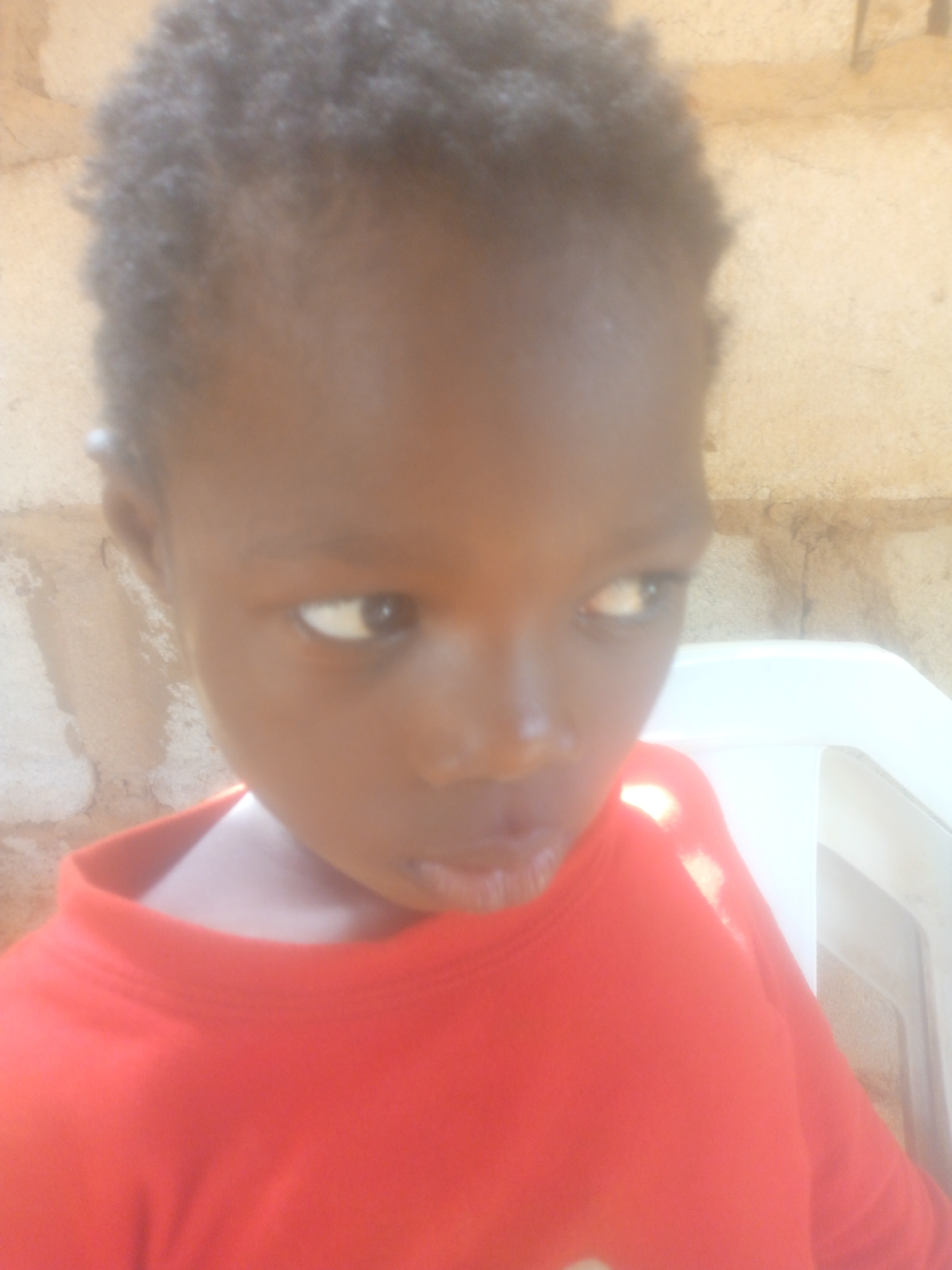 A close-up, soft-focus portrait features a young child, likely a toddler or preschooler, with dark skin and short, curly black hair. The child is looking towards the right of the frame with their eyes slightly averted, creating a pensive or curious expression. Their mouth is slightly open. They are wearing a plain red long-sleeved shirt with a ribbed collar.
The background consists of a textured wall, appearing to be light brown or tan, possibly made of brick or plaster. To the right of the child, a portion of a white plastic chair is visible. The lighting is soft and natural, suggesting it is daytime. There are no discernible activities or specific events taking place beyond the child's gaze. No text is visible in the image.