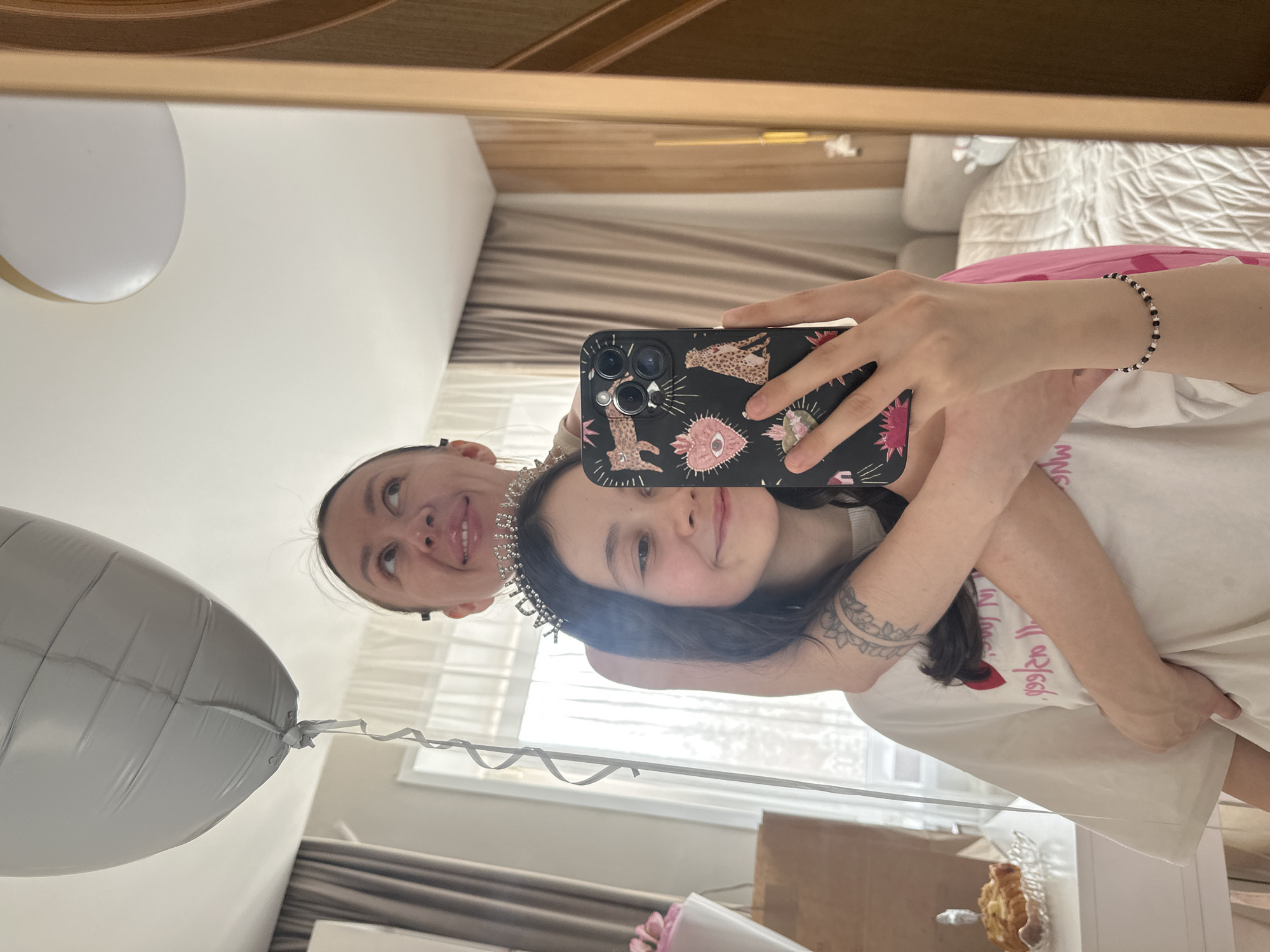 A mirrored selfie shows two women and a birthday celebration. The younger woman, with dark hair and wearing a tiara reading "Birthday," is holding a phone with a patterned case. She's wearing a white t-shirt and has a tattoo on her arm. The older woman stands behind her. A silver balloon is visible to the left. A bouquet and a small food item in a dish sit on a surface below. The room has a window, light curtains, and wood furniture. The location is not specified.