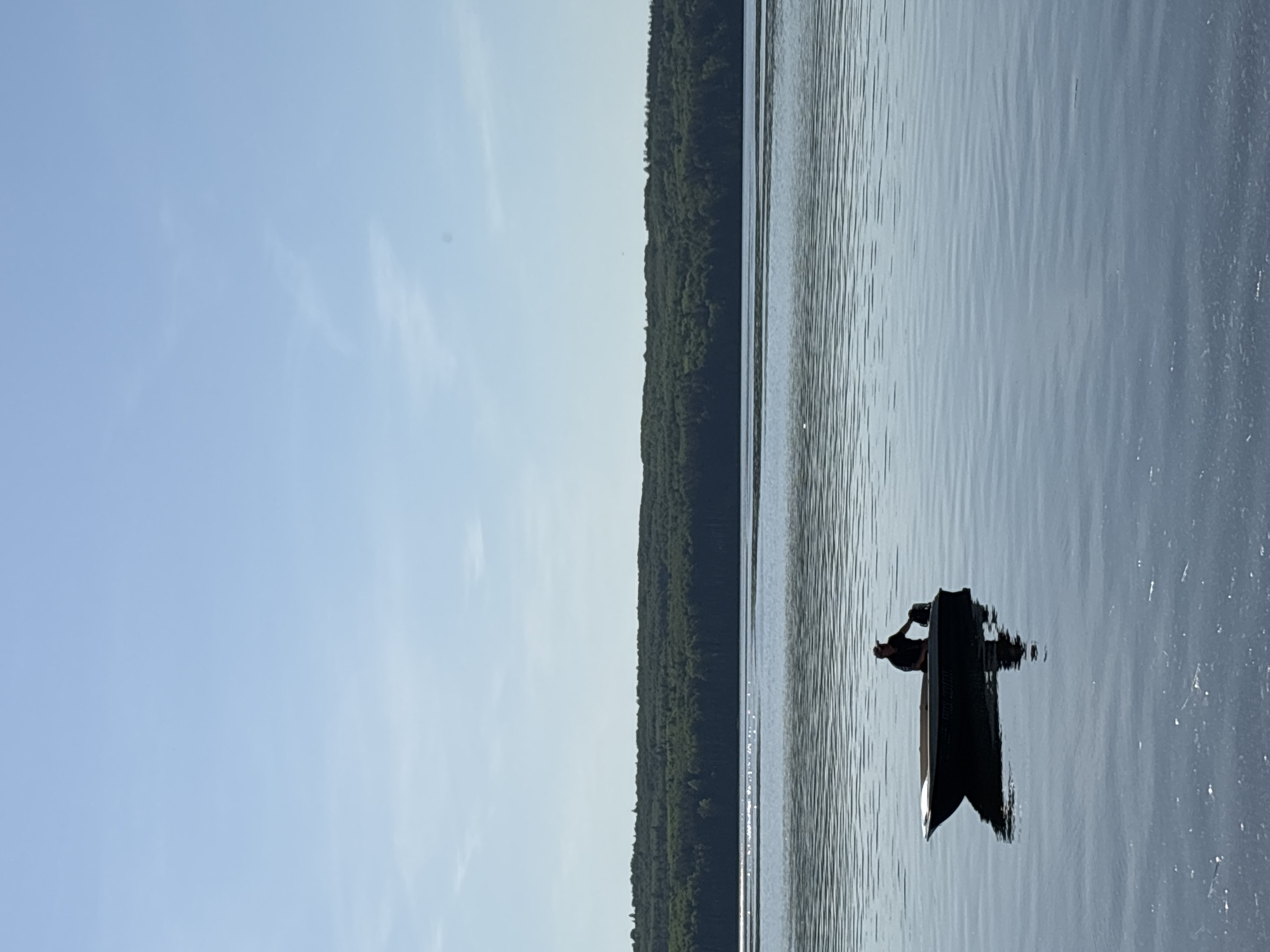 A landscape scene depicts a calm body of water, likely a lake or wide river, adjacent to a dense green forest. A single black boat or kayak sits on the water. A human figure is visible in the boat. The sky is blue with few clouds. This photo appears to be taken in Chaikovsky, Russia.