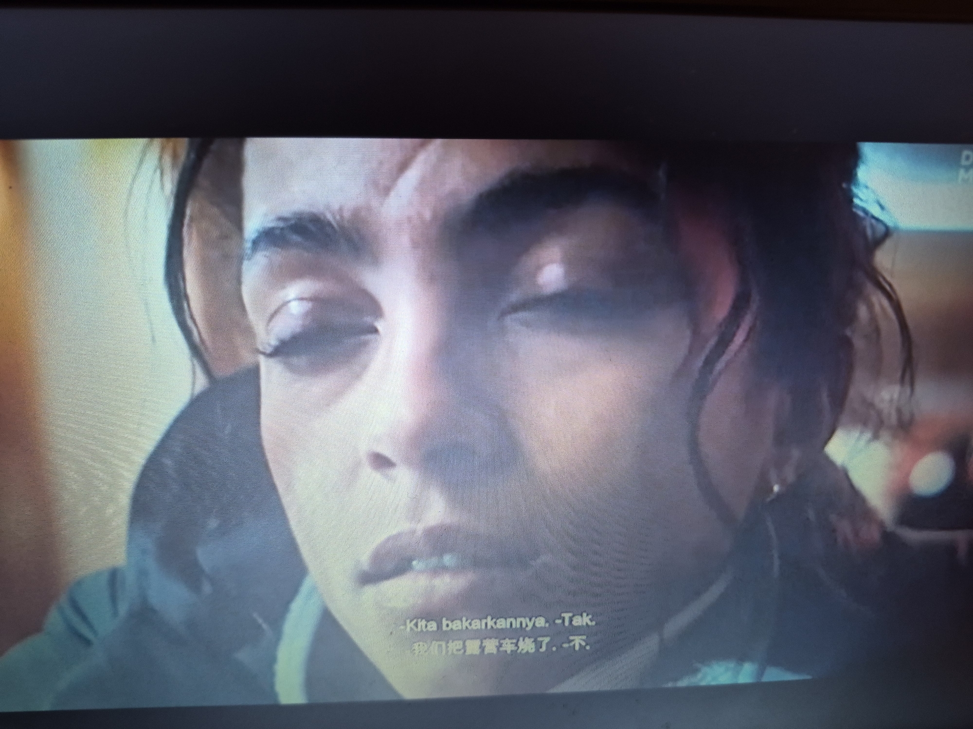 The image is a close-up of a person's face, primarily focusing on their eyes and mouth. The person has dark, curly hair and appears to be speaking or reacting to something. There are subtitles visible at the bottom of the frame in what appears to be Malay and Chinese characters, suggesting the scene is from a film or television show. The lighting is dim and somewhat diffused, with some areas of the face highlighted and others in shadow. The background is out of focus, showing indistinct shapes and colors that do not provide specific environmental clues. There is no discernible information within the image that would allow for the identification of a specific location in the city of Sandwell, United Kingdom.