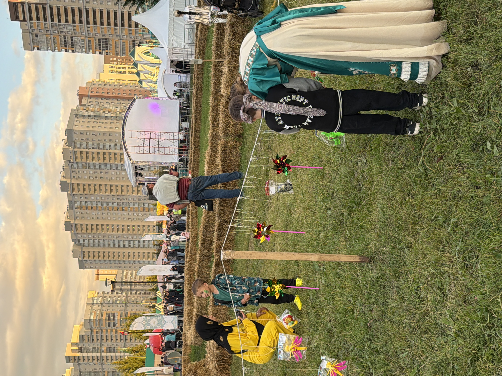 This is a full-shot, eye-level, outdoor medium-distance photograph of an event. In the foreground, two children are standing in a grassy area with hay bales behind them. The child on the left is wearing a yellow hijab and outfit, and is looking down and to the left, appearing to be interacting with the child next to them. The child on the right is wearing a green shirt and a dark patterned jacket, and is looking to their right. Between the children, a wooden pole is planted in the ground, from which multiple colorful pinwheels and toys are hanging on strings. In the background, several people are gathered, and large buildings and tents are visible, suggesting an urban setting. The sky is cloudy and appears to be at sunset.