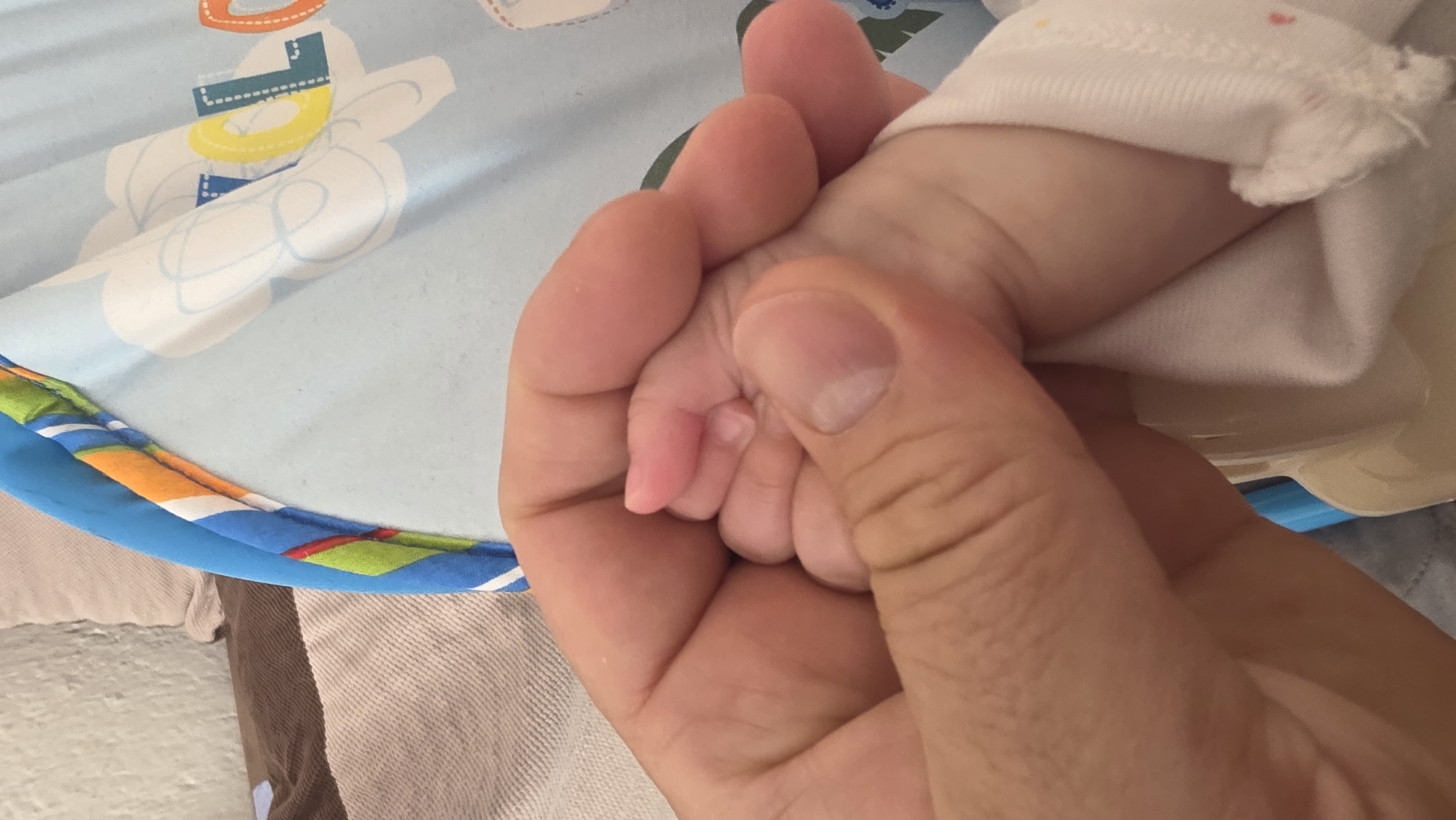 A close-up shot shows an adult hand holding a baby's hand. The baby is wearing a white garment. The background includes a light blue surface with colorful patterns. The setting is indoors. The city of Blagoevgrad, Bulgaria is not identifiable in the image.