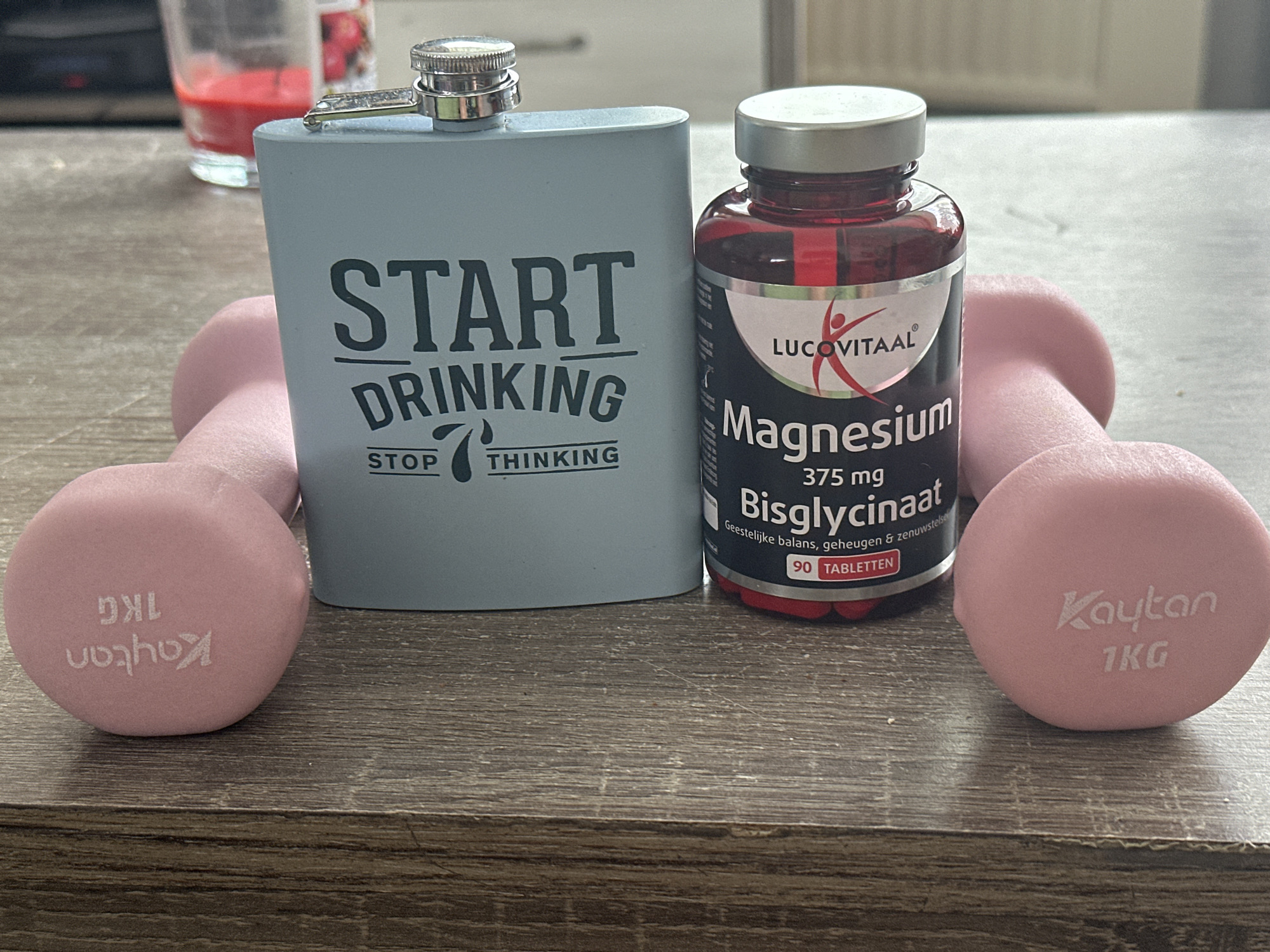 This image depicts a still life arrangement on a wooden surface, possibly a table. The primary objects are a light blue hip flask with "START DRINKING STOP THINKING" printed on it in black lettering, a bottle of "LUCOVITAAL Magnesium Bisglycinaat 375 mg" containing 90 tablets, and two pink dumbbells, each weighing 1KG and branded "Kaytan". In the background, out of focus, are two glasses, one with a red liquid. The composition suggests a juxtaposition of leisure and health. The lighting is natural, casting soft shadows. The scene is indoors, with the background hinting at a domestic setting. The specific location in Steenwijk, Netherlands, cannot be determined from the visual information alone.