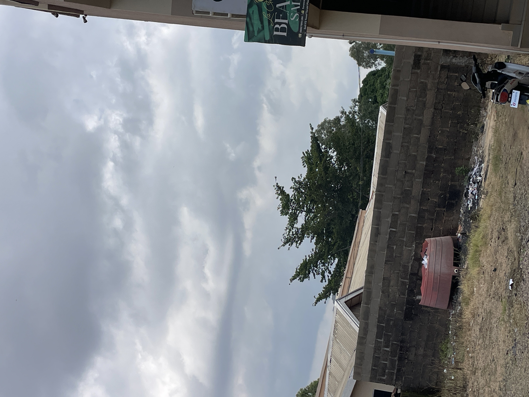 A vertical, outdoor shot shows a cloudy sky with a building in the foreground and some trees in the background. A sign hangs from the building, reading "BEAUTY SALON" and phone numbers. To the right, a red barrel sits on the ground next to a brick wall. A motorcycle is parked near the barrel. The ground is covered with dry grass and some litter. The scene suggests a casual, everyday setting in Jos, Nigeria, possibly in the morning or late afternoon given the cloudy weather.