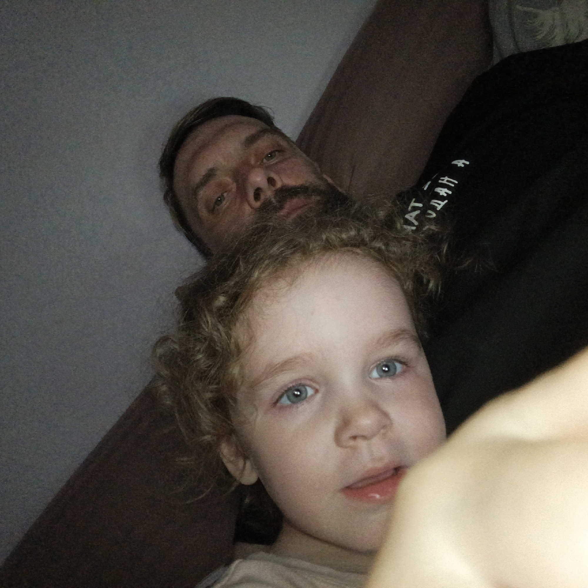 The image captures two individuals, an adult male and a young child, in a selfie-style photograph, purportedly located in the city of Марковский, Russia. The adult male is positioned in the background, partially obscured by the child, with his head oriented towards the upper left. He possesses a dark beard and mustache, dark hair, and light-colored eyes. His expression is neutral. He is wearing a dark top, likely a t-shirt or hoodie, with fragmented white text visible on the left shoulder area, which reads "АТ _ДАН А," potentially Cyrillic characters mirrored from a selfie perspective.
In the foreground, a young child with curly, light-colored hair and prominent blue eyes is positioned in the lower central and right portions of the frame. Her mouth is slightly open, revealing a pink lower lip. She wears a light-colored garment. A blurry, light-toned object, consistent with a human limb or hand, partially obstructs the right side of the child's face. The background is dim and indistinct, featuring a lighter gray surface on the upper left and a darker brown, possibly fabric, surface on the upper right, suggesting an indoor setting. Both individuals are looking towards the camera.