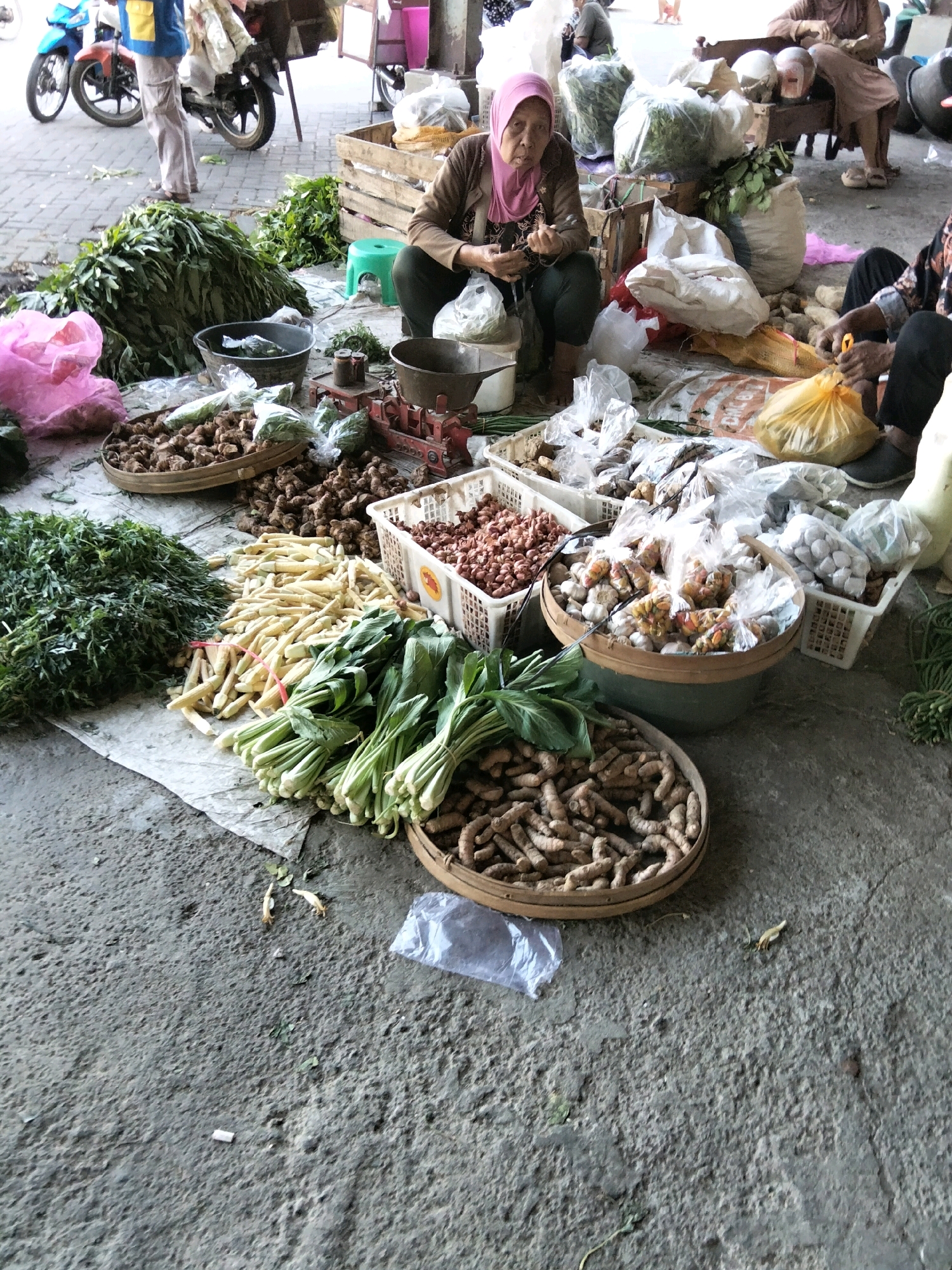 The image depicts an outdoor market scene, likely in Banyudono, Indonesia, given the context. In the foreground, various fresh produce is displayed on a concrete ground covered with a patterned cloth. Piles of turmeric roots in a woven basket and loose on the ground are prominent. To the left, large bunches of leafy greens are stacked, along with smaller bundles of what appear to be baby corn or similar vegetables. Further back, in a large woven basket, are more turmeric roots. Several white plastic baskets contain an assortment of goods, including smaller vegetables like shallots, garlic, and what seem to be small dried chilies. Some items are bagged in clear plastic. In the middle ground, a woman wearing a pink hijab and a brown top is seated, interacting with items in front of her and looking towards the camera. Another person, partially visible, is seated to the right, engaged in an activity with a yellow plastic bag. In the background, a few motorcycles are parked, and other market stalls or activities can be inferred. The overall environment suggests a bustling local marketplace with an emphasis on fresh produce.