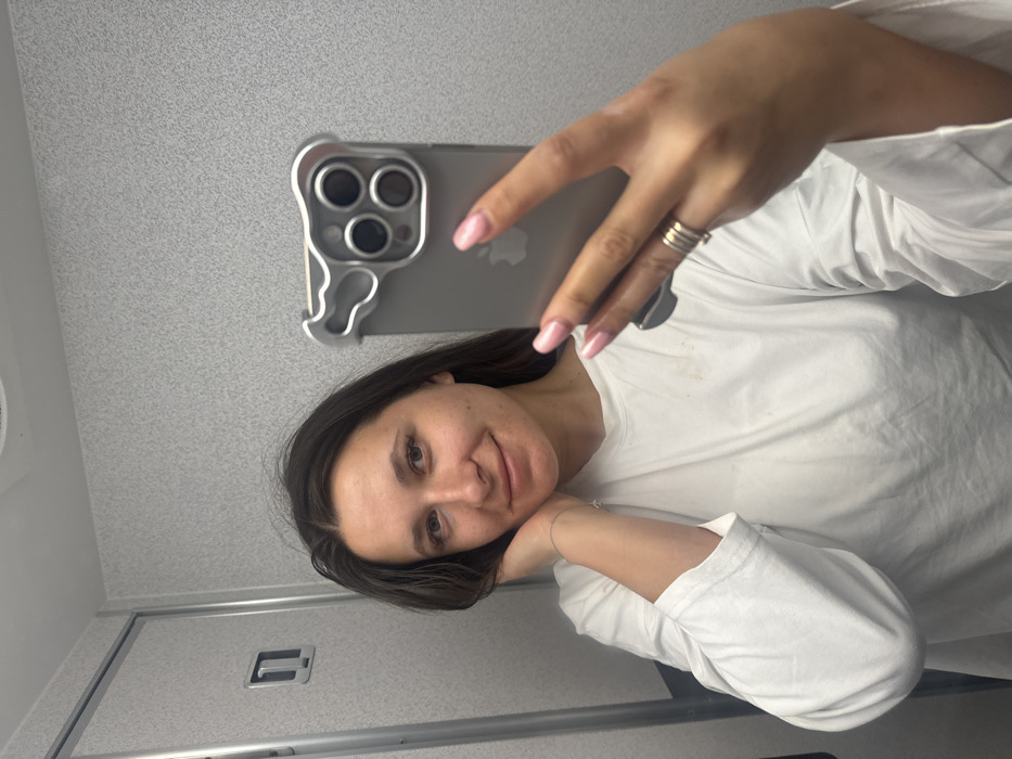 The image captures a close-up mirror selfie of a young woman, appearing to be in an enclosed space such as an airplane lavatory. The woman is smiling gently, with her head tilted slightly, and her left hand resting casually under her chin. She has long, dark, straight hair parted slightly off-center, and light-colored eyes. She is wearing a simple white, long-sleeved top. Her fingernails are manicured with light pink polish, and she wears a silver-colored ring with multiple bands on the ring finger of her right hand, and a thin silver-colored bracelet on her left wrist.
She is holding a silver-colored iPhone, likely a recent model given the large triple camera array, which is encased in a distinctive silver, possibly metallic, protective case. The case has a unique, somewhat ergonomic shape around the camera module, and the Apple logo is visible through the case. Her right hand, holding the phone, is prominently featured in the upper central part of the image.
The background, reflected in the mirror, consists of light gray, speckled, utilitarian panels on what appear to be walls and a ceiling. In the lower left portion of the image, a simple silver, horizontal door handle is visible, further suggesting a small, functional room. The lighting is even and bright, consistent with artificial indoor lighting. The overall image is rotated 90 degrees counter-clockwise from a standard portrait orientation.