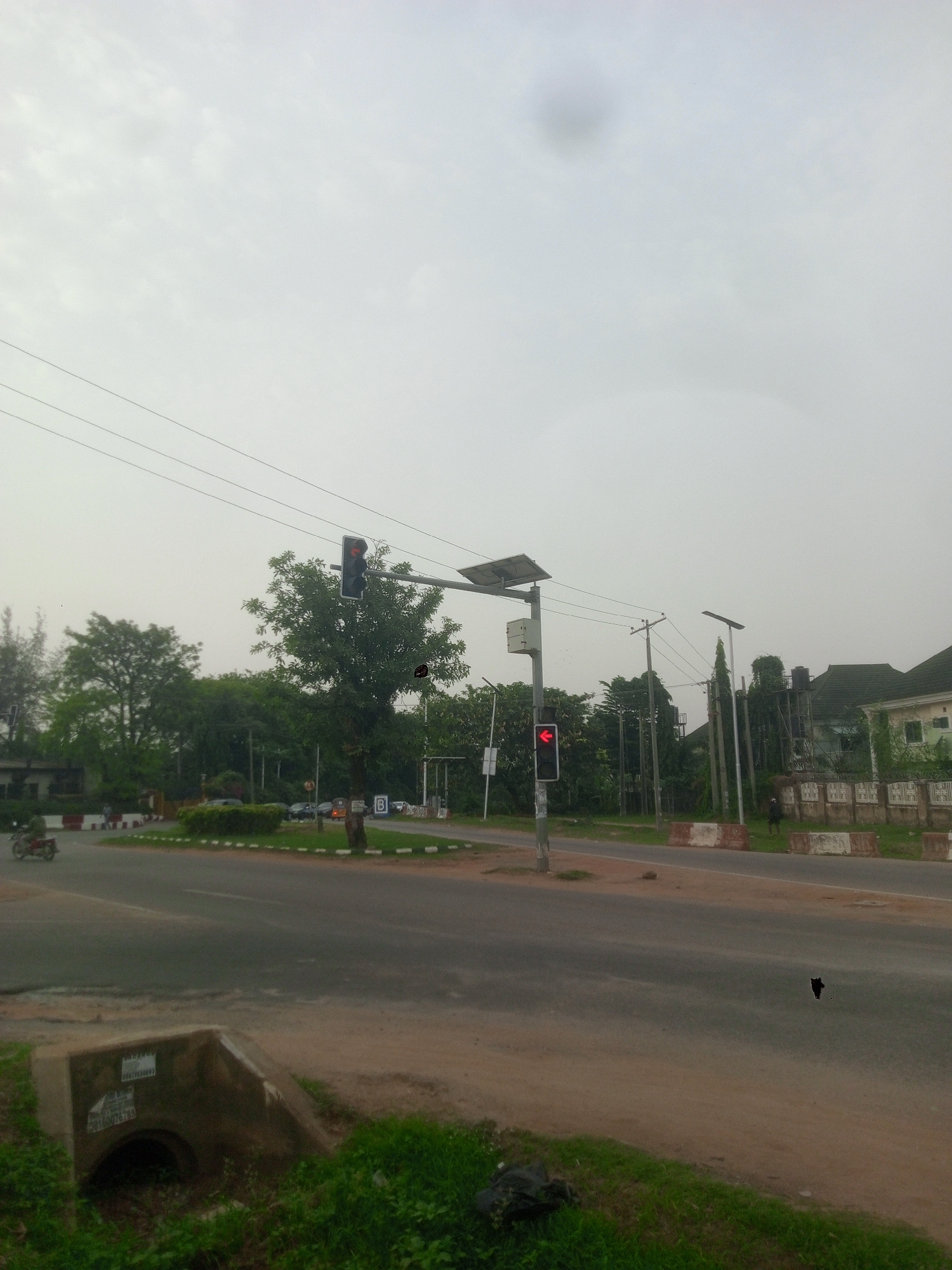 The image shows a street intersection in Gwarinpa, Nigeria on an overcast day. Two sets of traffic lights are visible, one showing a red light and the other showing the red light for straight ahead. A roundabout with a small patch of greenery sits at the intersection. There are trees, buildings, utility poles, and overhead wires around the intersection. A motorcycle is near the roundabout. A concrete drainage culvert is in the foreground. A pedestrian is by the side of the road. The road surface appears worn. There is trash on the road.