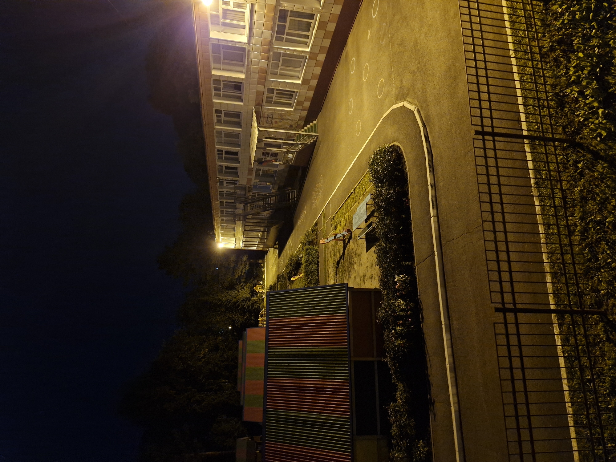 An exterior night-time view of a playground area next to a two-story building. The building has a light-colored facade with numerous windows and a covered entrance with stairs. To the left of the building is a paved area with a curved walkway and a black metal fence in the foreground. On the playground, there is a brightly colored playhouse with a striped roof and another smaller structure with a striped roof in the background. A seesaw and a blue bench are also visible. The scene is illuminated by artificial lighting, creating long shadows. The location is identified as Kemerovo, Russia.