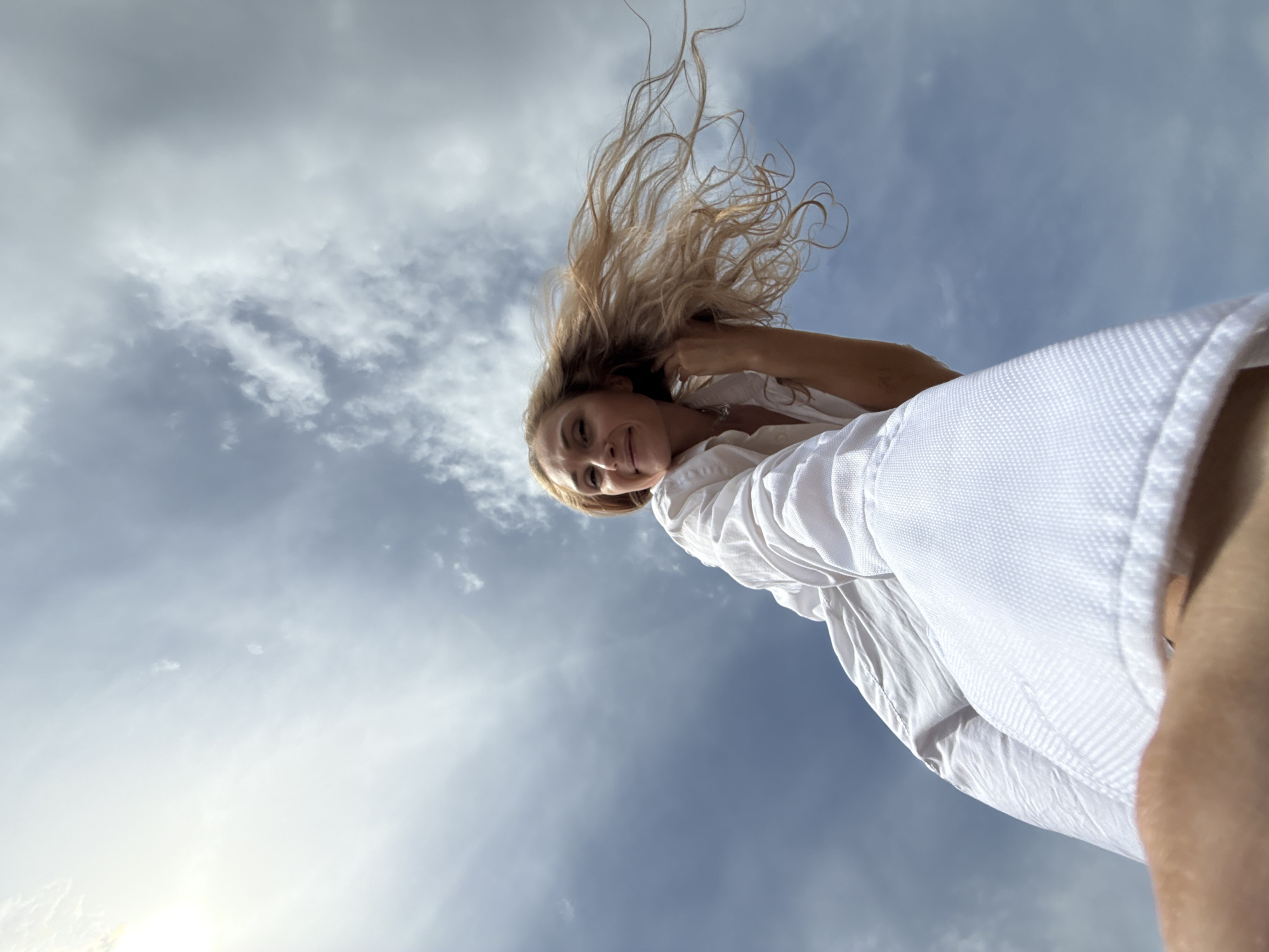 A low-angle, upward-facing shot shows a woman with blonde hair against a backdrop of a sky with scattered clouds. The woman's hair is windswept, some strands appearing to be in motion. She is wearing a white, textured shirt. Her expression is a slight smile. The sky is a mix of blue and white, with sunlight visible near the bottom left corner of the frame. The location is not discernible from the image itself. No information can be provided to verify if this image was taken in the city of Chaikovsky, Russia.